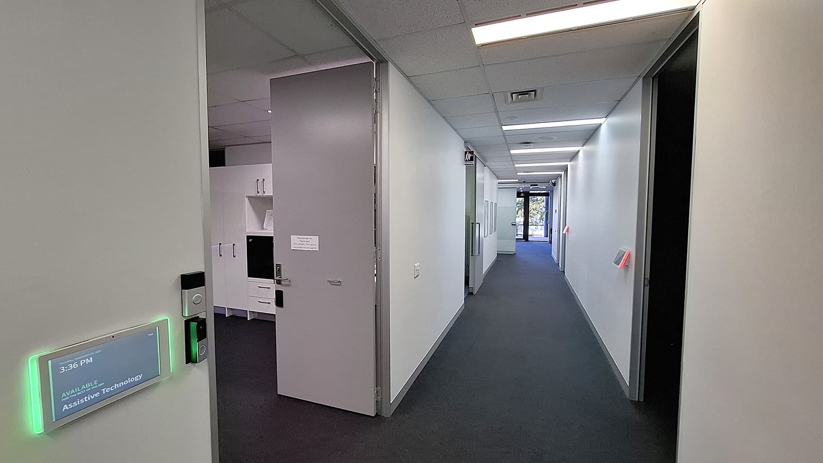 On the left, a large television screen mounted on the wall above a display table with a model labelled "Spinal Life Healthy Living Centre" and a potted plant beside it. In the middle, a long hallway with white walls, a grey carpet, and fluorescent ceiling lights, leading to a glass door at the end. On the right, a white reception desk with a computer monitor and informational posters on the wall. The ceiling is white with recessed lighting.