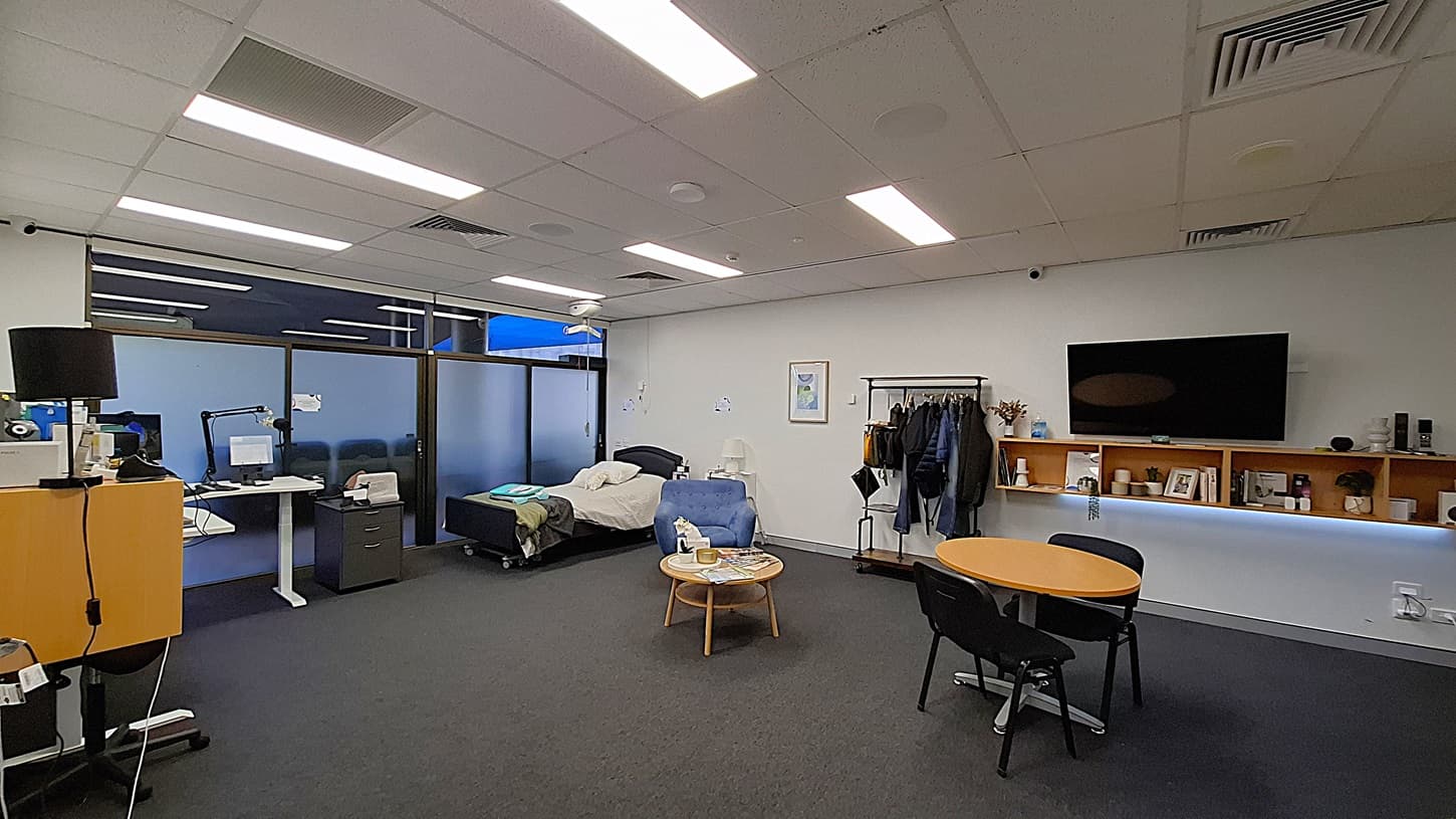 On the left, a desk with a computer monitor, a black lamp, and a filing cabinet. In the middle, a bed with white sheets and a blue blanket, a small table with a tissue box and a water bottle, and a blue armchair. On the right, a wall-mounted television above wooden shelves holding various items, and a coat rack with jackets. The floor is dark grey carpet, and the ceiling has white panels with fluorescent lighting.