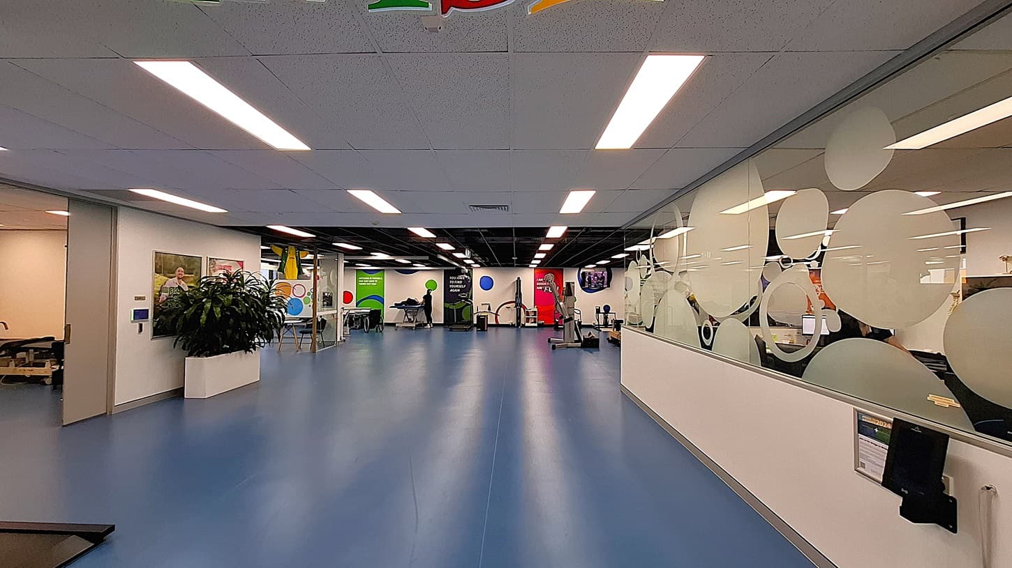 On the left, an open area with blue flooring, exercise equipment, and colourful wall decorations. In the middle, a glass partition with frosted circular designs and a "Finish Line" banner hanging above. On the right, a hallway with dark carpet, a white shelving unit holding various items and a potted plant, and a black bin. The ceiling has white panels with recessed lighting and air vents.