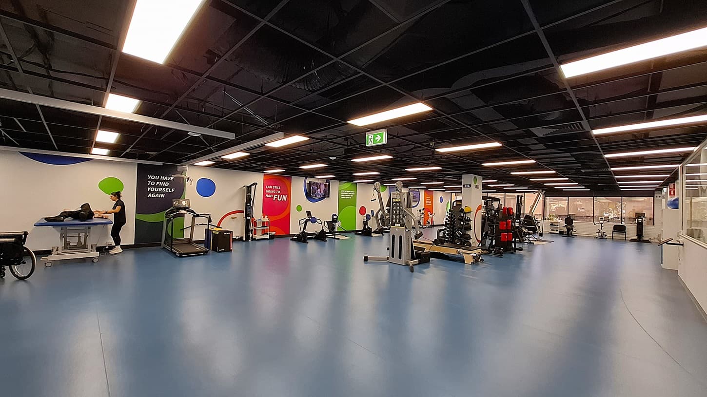 On the left, a person working on a black exercise table next to a wall with motivational text and colourful circular designs. In the middle, various exercise machines including treadmills and stationary bikes, with bright wall graphics. On the right, more exercise equipment, weight racks, and a glass partition with a view of an office area. The floor is blue, and the ceiling is black with exposed structures and recessed lighting.