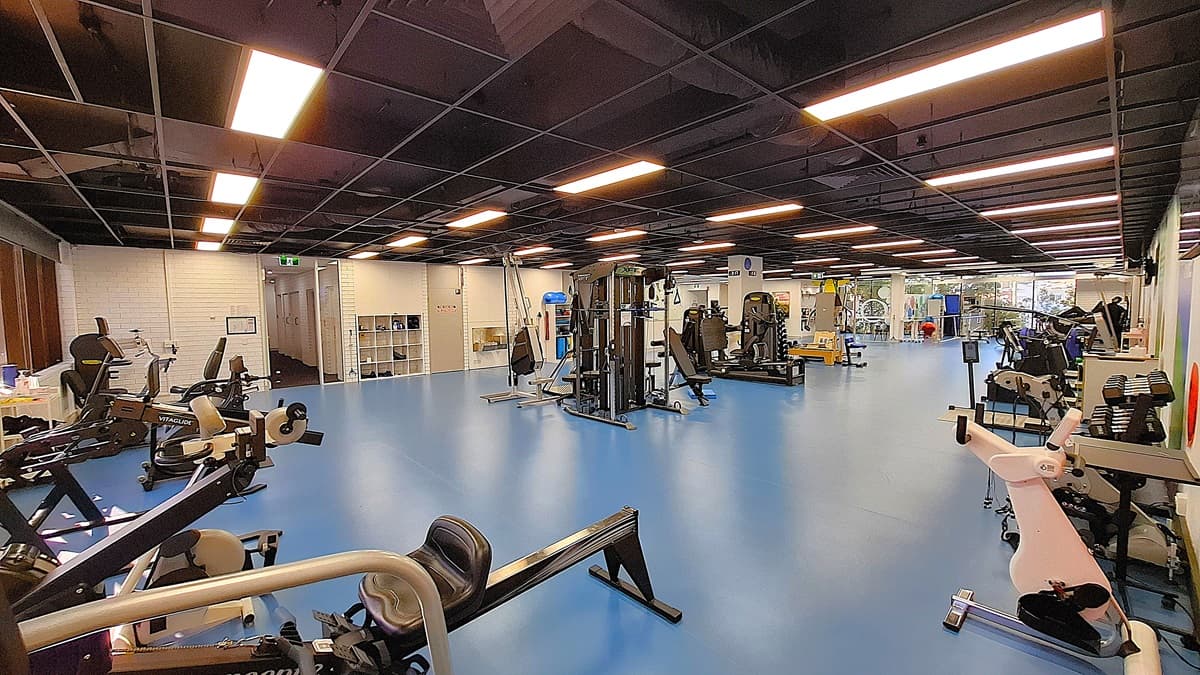 On the left, various exercise machines including stationary bikes and rowing machines, positioned near a white brick wall with a small window. In the middle, a spacious area with blue flooring and multiple weight and resistance training machines. On the right, more exercise equipment including treadmills and weight racks, with a large window in the background. The ceiling is black with exposed structures and evenly spaced fluorescent lighting.