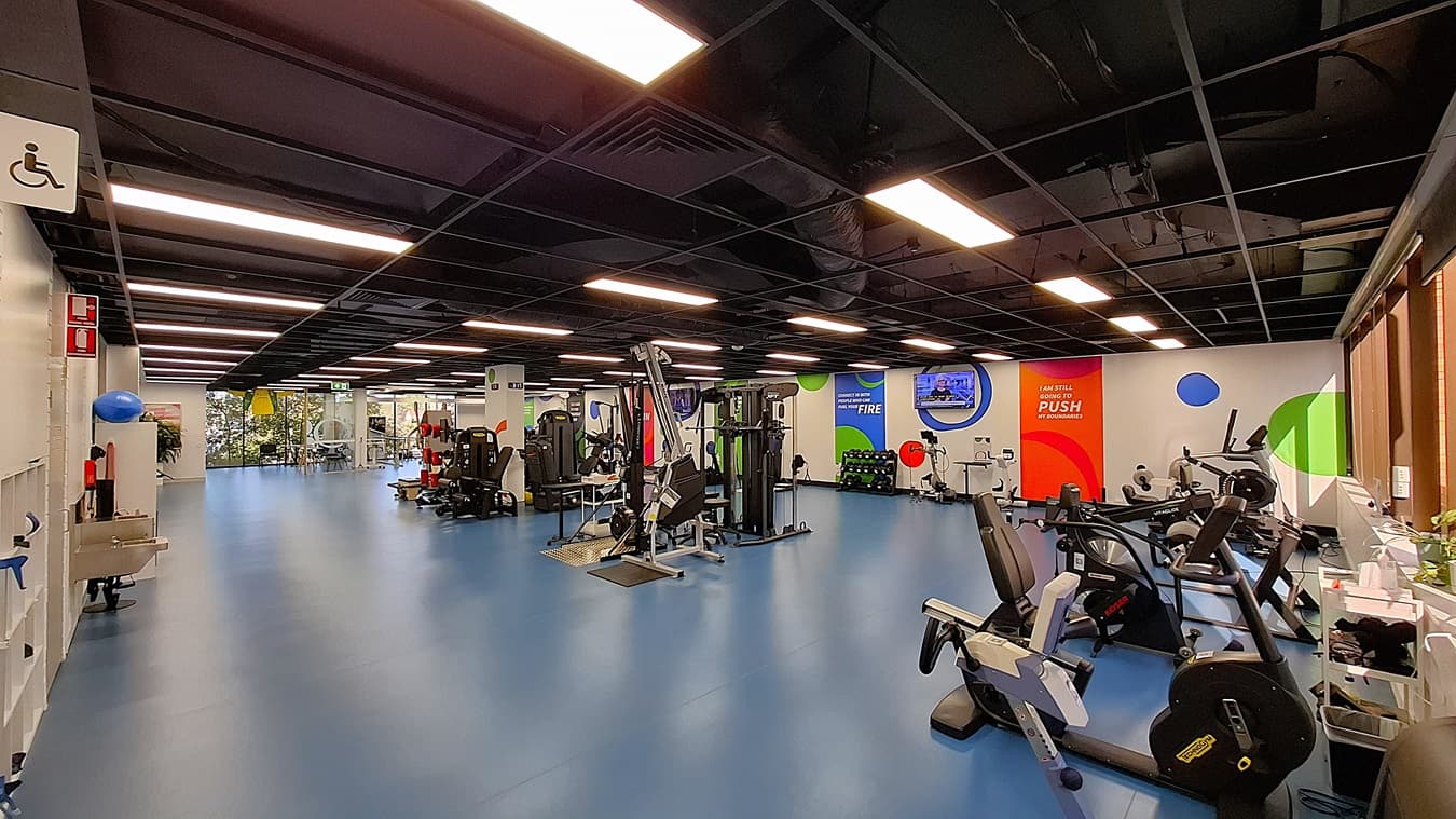 On the left, a white wall with a sink, shelves holding exercise balls, and a fire extinguisher. In the middle, a spacious area with blue flooring, various exercise machines including weight and resistance training equipment, and colourful motivational banners on the walls. On the right, more exercise equipment including stationary bikes and treadmills, with large windows letting in natural light. The ceiling is black with exposed structures and evenly spaced fluorescent lighting.
