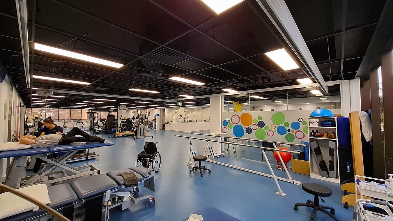 On the left, a person working on a blue treatment table next to a wall with colourful circular designs. In the middle, a spacious area with blue flooring, various exercise machines, and a wheelchair. On the right, a white shelving unit holding exercise balls and equipment, and a parallel bar setup for rehabilitation. The ceiling is black with exposed structures and evenly spaced fluorescent lighting.