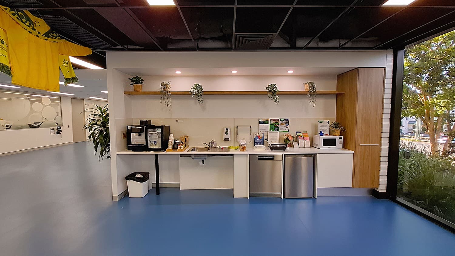 On the left, a hallway with blue flooring, a large potted plant, and a wall with frosted circular designs. In the middle, a kitchenette with a coffee machine, sink, microwave, and informational brochures on the counter. Above the counter, a wooden shelf with potted plants. On the right, a large window with a view of greenery outside. The floor is blue, and the ceiling is black with exposed structures and recessed lighting.