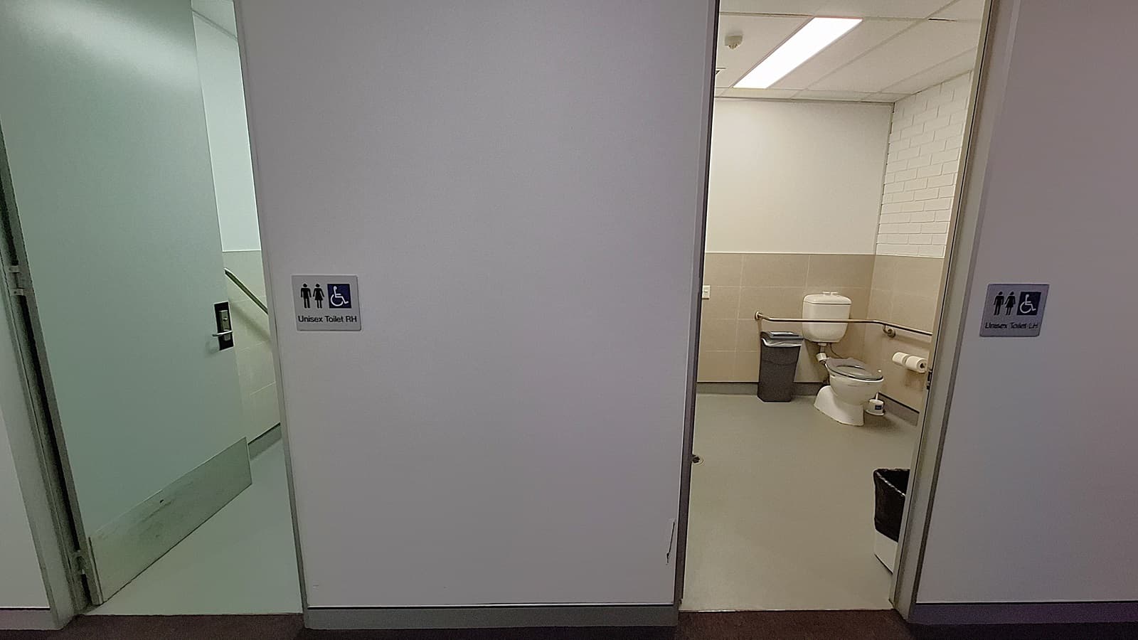 On the left, an open door leading to a stairwell with a handrail and a sign indicating a unisex accessible toilet. In the middle, a white wall separating two doorways. On the right, an open door revealing a bathroom with a toilet, grab bars, a waste bin, and a toilet paper holder. The floor is grey, and the ceiling has white panels with fluorescent lighting.