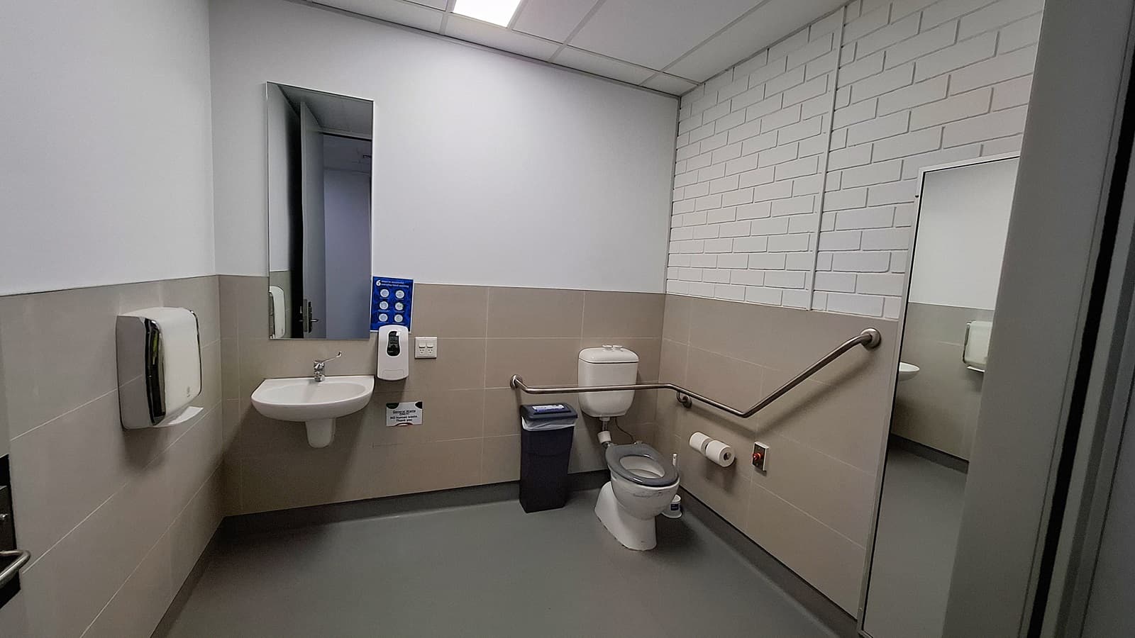 On the left, a white sink with a mirror above it, a soap dispenser, and a paper towel dispenser. In the middle, a grey waste bin and a white toilet with a grab bar on the right side. On the right, a large mirror reflecting the sink area and a toilet paper holder mounted on the wall. The floor is grey, and the ceiling has white panels with fluorescent lighting. The walls are half-tiled in beige with the upper half painted white, and one wall has white brickwork.