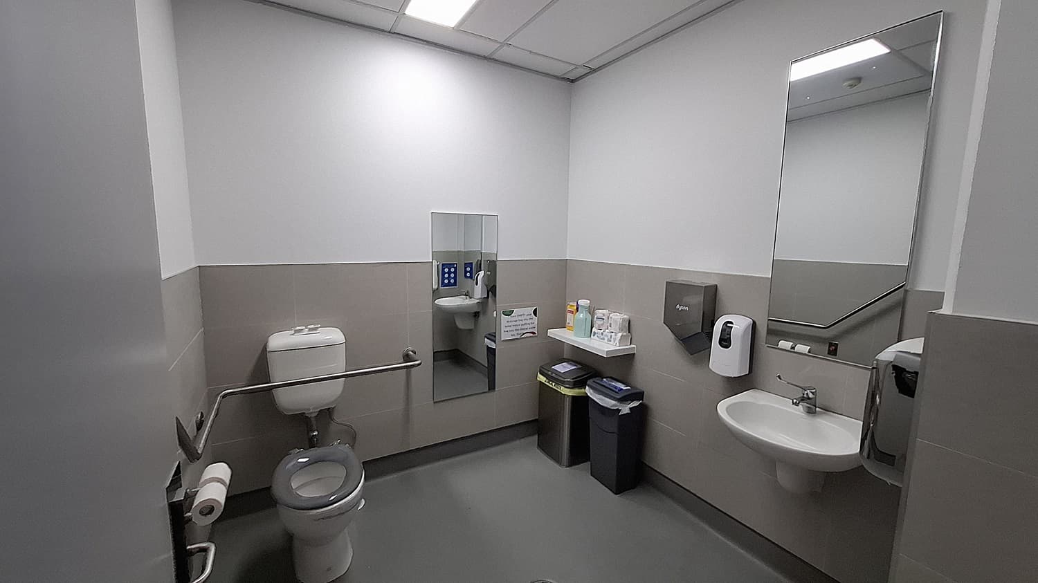 On the left, a white toilet with a grey seat, a grab bar, and a toilet paper holder. In the middle, a wall-mounted mirror above a shelf holding cleaning supplies, with two bins below. On the right, a white sink with a soap dispenser, a paper towel dispenser, and a large mirror above. The floor is grey, and the ceiling has white panels with fluorescent lighting. The walls are half-tiled in beige with the upper half painted white.