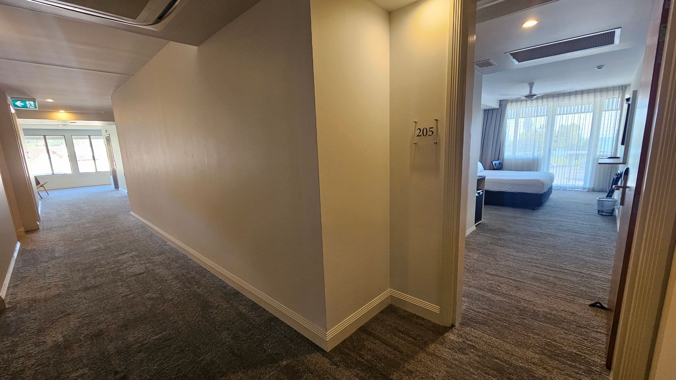 A hotel corridor with a beige carpet and cream walls. On the right, a door marked "205" is open, revealing a room with a large bed, a chair, and sheer curtains covering large windows. The ceiling has recessed lighting and an air conditioning vent. On the left, the corridor extends towards a window with a view of greenery, and an emergency exit sign is visible.