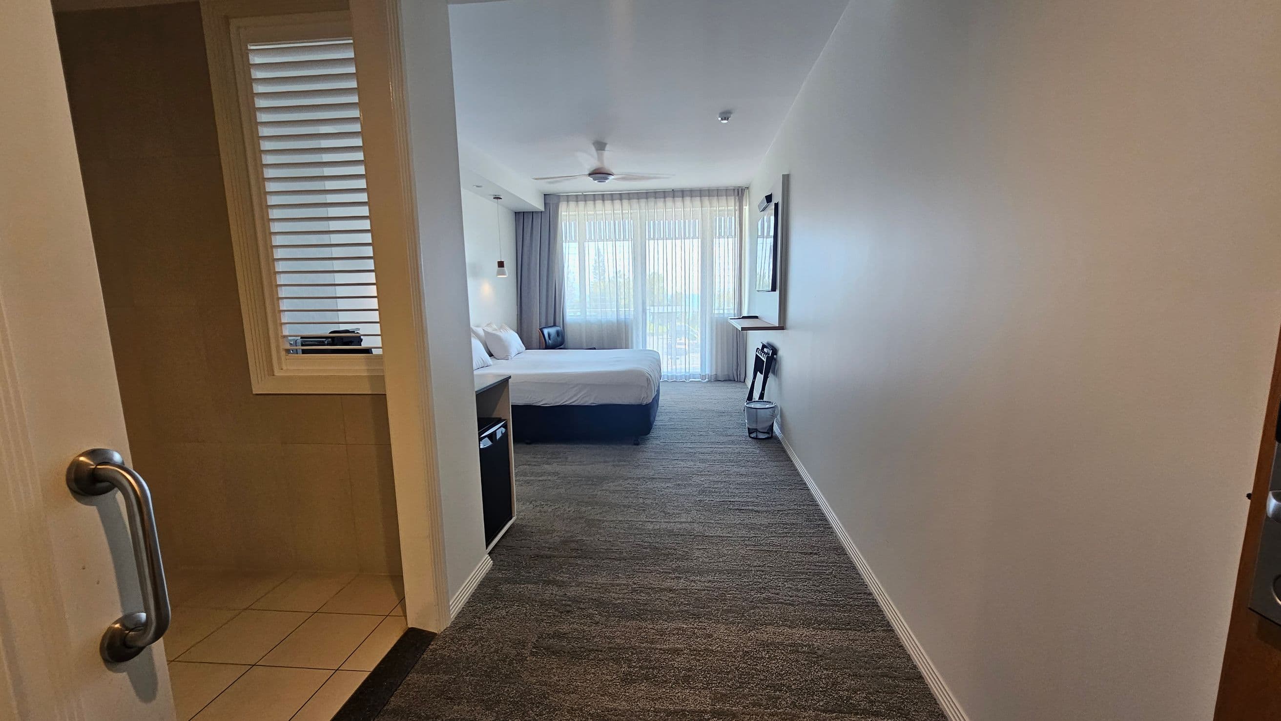 A hotel room with a view from the entrance. On the left, there's a partially open door with a metal handle leading to a tiled area. Further in, a bed with white linens is against the left wall, accompanied by a bedside table and lamp. On the right, a wall-mounted TV and a folded luggage rack are visible. The room has a carpeted floor and a ceiling fan. At the far end, sheer curtains cover a large window or balcony door, allowing natural light to enter.