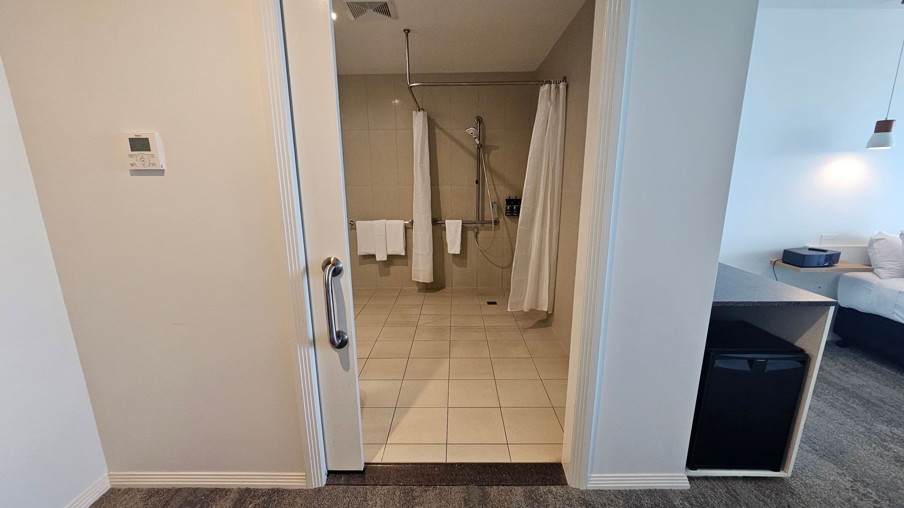 A bathroom with a tiled floor and a walk-in shower featuring a curtain and a handheld showerhead. Towels hang on a rail. On the left wall, there's a thermostat. The room outside has a carpeted floor, a small fridge under a counter, and a bed with white bedding. A pendant light hangs above the bed.