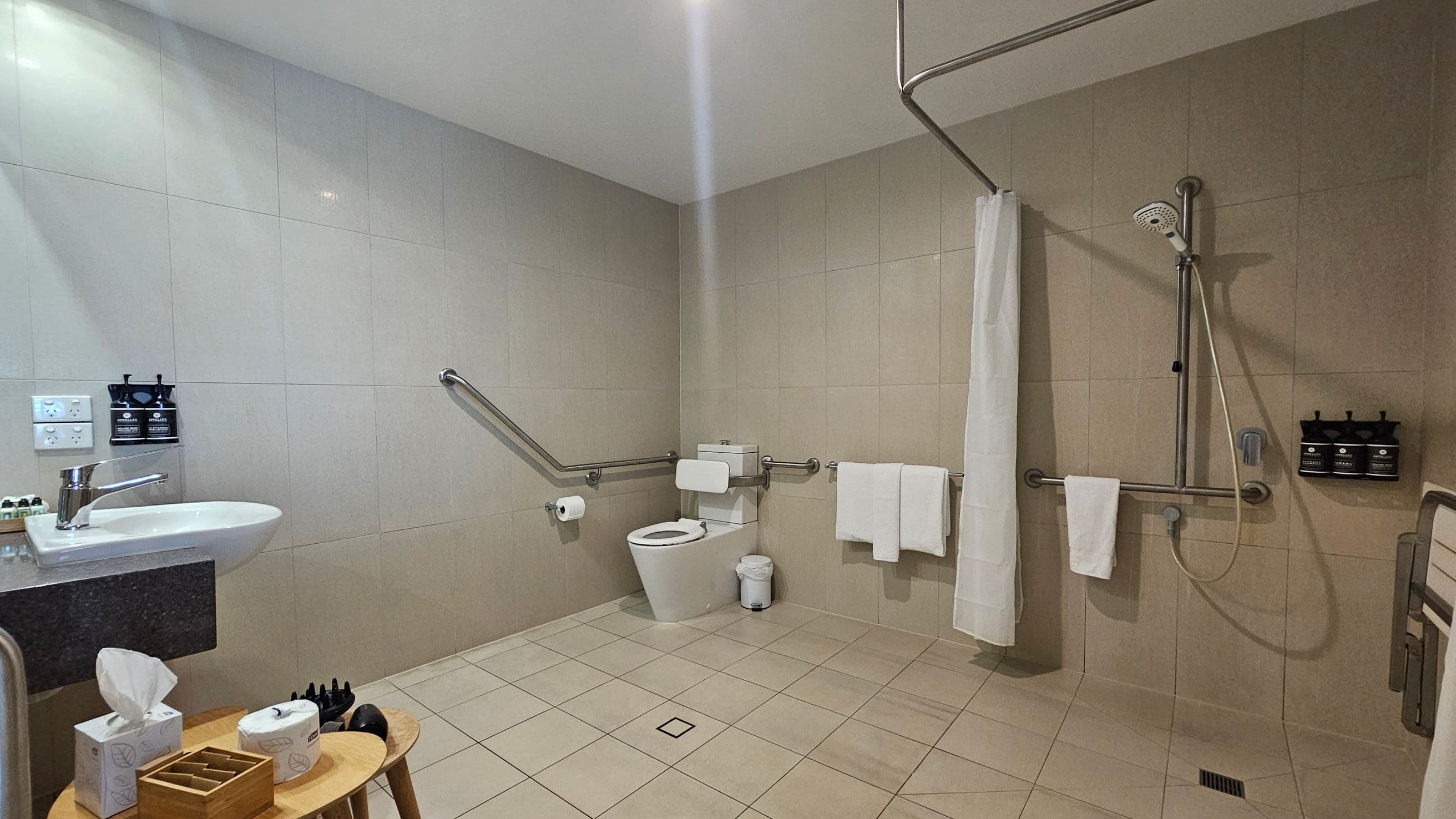 A spacious bathroom with beige tiled walls and floor. On the left, a white sink with a silver tap is mounted on a dark countertop, with soap dispensers and a light switch nearby. Below, a wooden stool holds a tissue box and toiletries. In the centre, a toilet with a support rail and a small bin. On the right, a walk-in shower with a handheld showerhead, a white curtain, and wall-mounted soap dispensers. Several grab bars and white towels are present throughout.