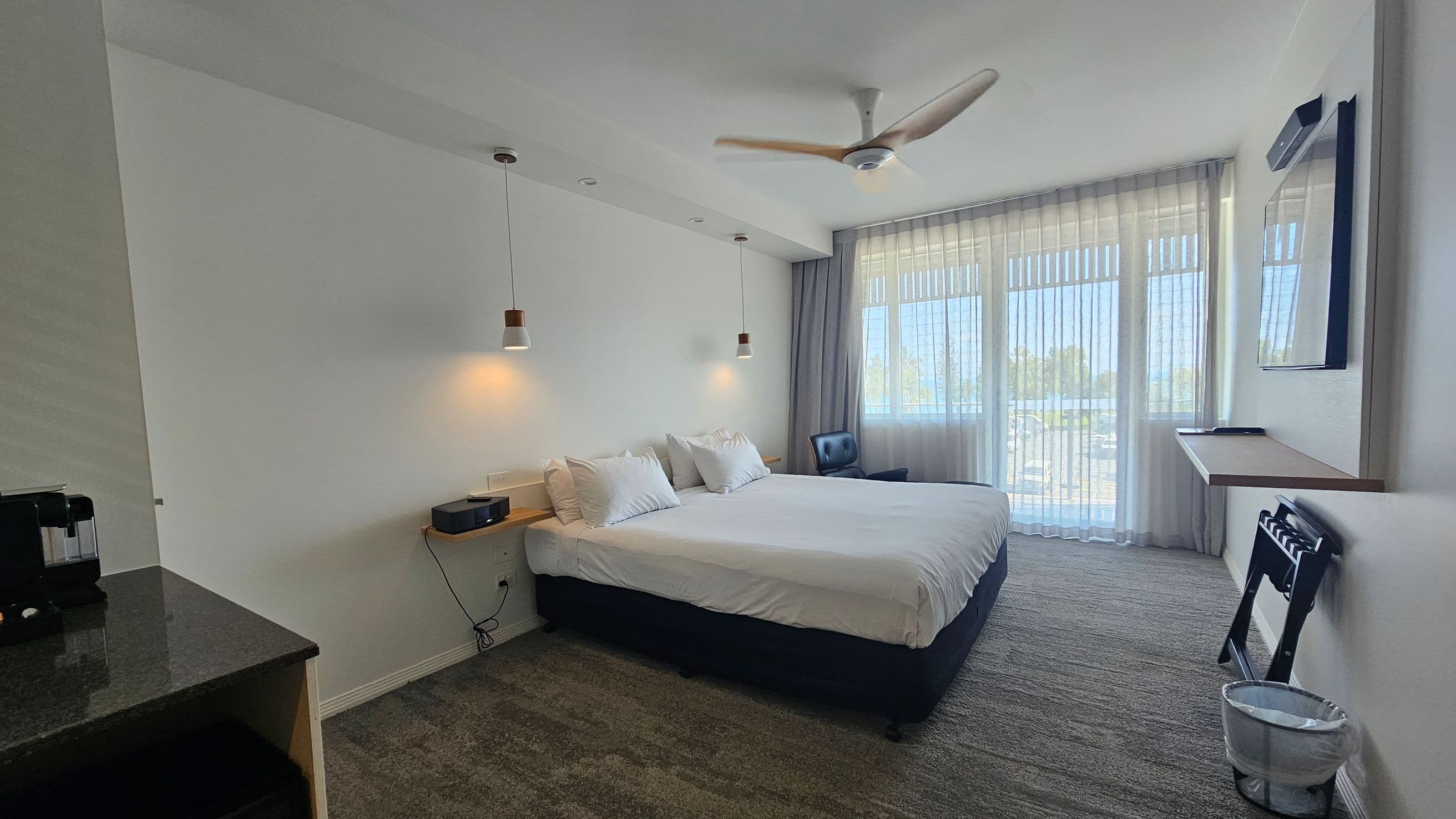 A modern hotel room with a large bed in the centre, adorned with white linens and pillows. Two pendant lights hang above the bed. On the left, a black countertop with a coffee machine. To the right, a window with sheer curtains allows natural light in, next to a blue chair. A wall-mounted TV and a small desk are on the right wall. The floor is carpeted in a mix of green and grey tones, and a ceiling fan is above. A small waste bin is on the floor near the desk.