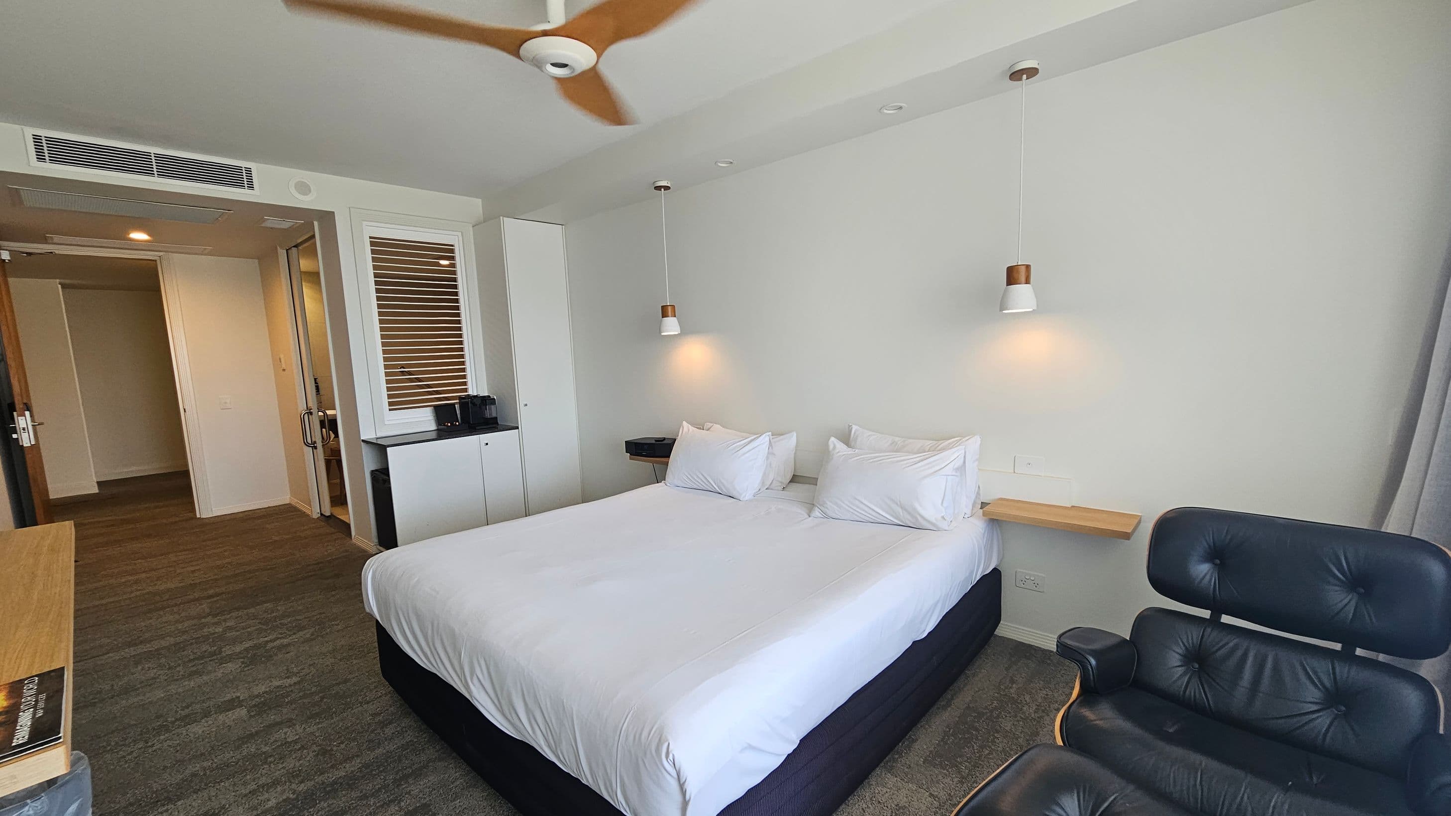 A modern hotel room with a large bed in the centre, adorned with white linens and pillows. Above the bed, two pendant lights hang from the ceiling. To the right, a black leather chair is positioned. On the left, a small cabinet with a coffee machine is placed beneath a window with blinds. The entrance to the room is visible in the background, with a wooden door and a glimpse of the bathroom. The floor is carpeted in a mix of grey and green tones, and a ceiling fan is overhead.