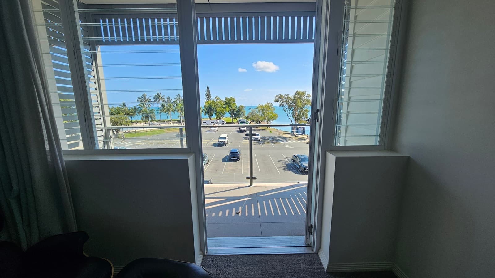 View through an open door with louvred windows on either side, leading to a balcony overlooking a car park. Beyond the car park, there are palm trees and a glimpse of the sea. The interior has a dark carpeted floor and light-coloured walls.
