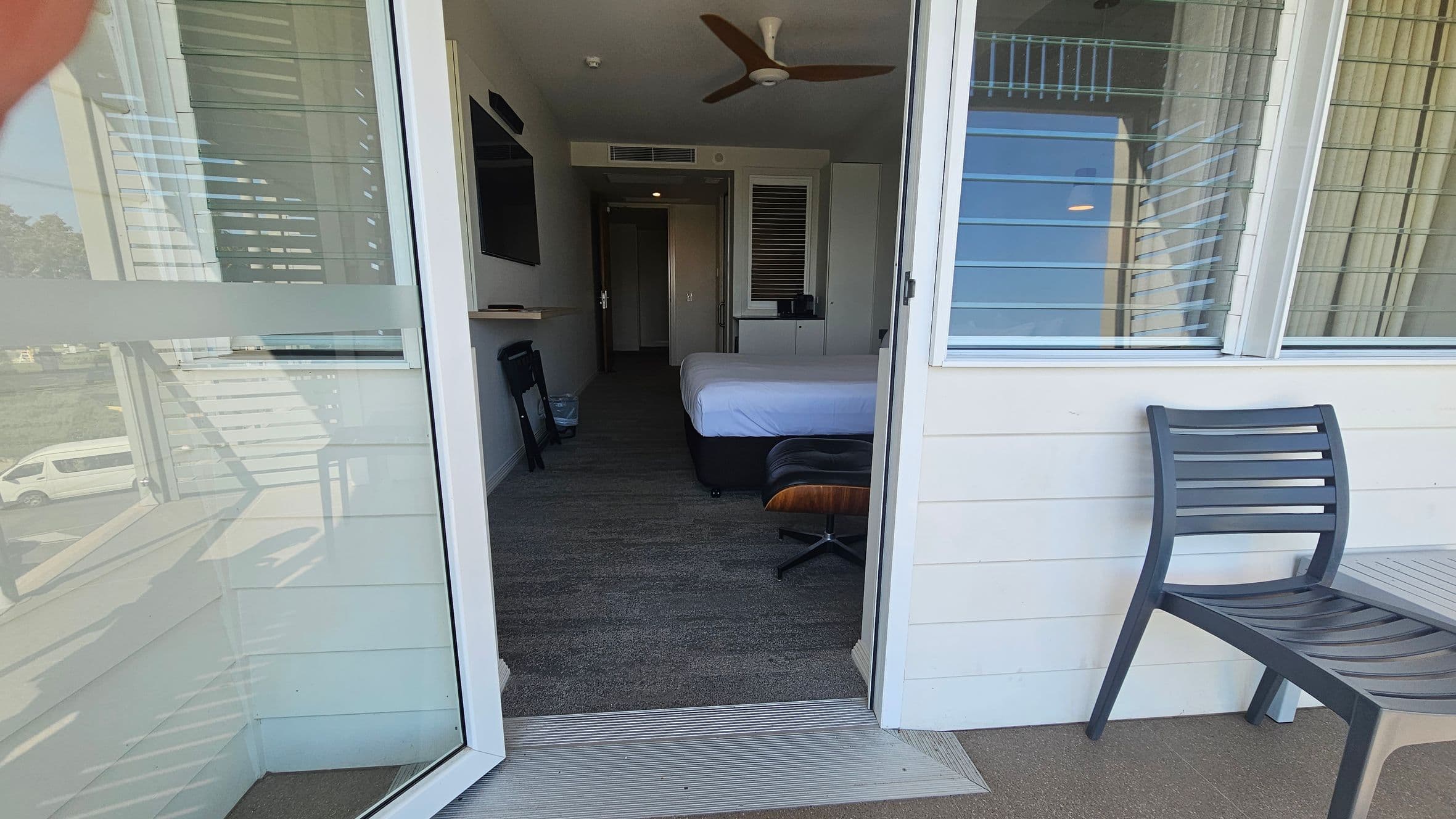 A view from a balcony into a hotel room. On the right, a dark grey chair is placed against the wall. The room has a bed with white sheets, a brown chair, and a flat-screen TV mounted on the left wall. A ceiling fan is visible above. The floor is carpeted, and there is a small table and a bin near the TV. The room has a window with horizontal blinds. Outside, a white van is parked on a grassy area.