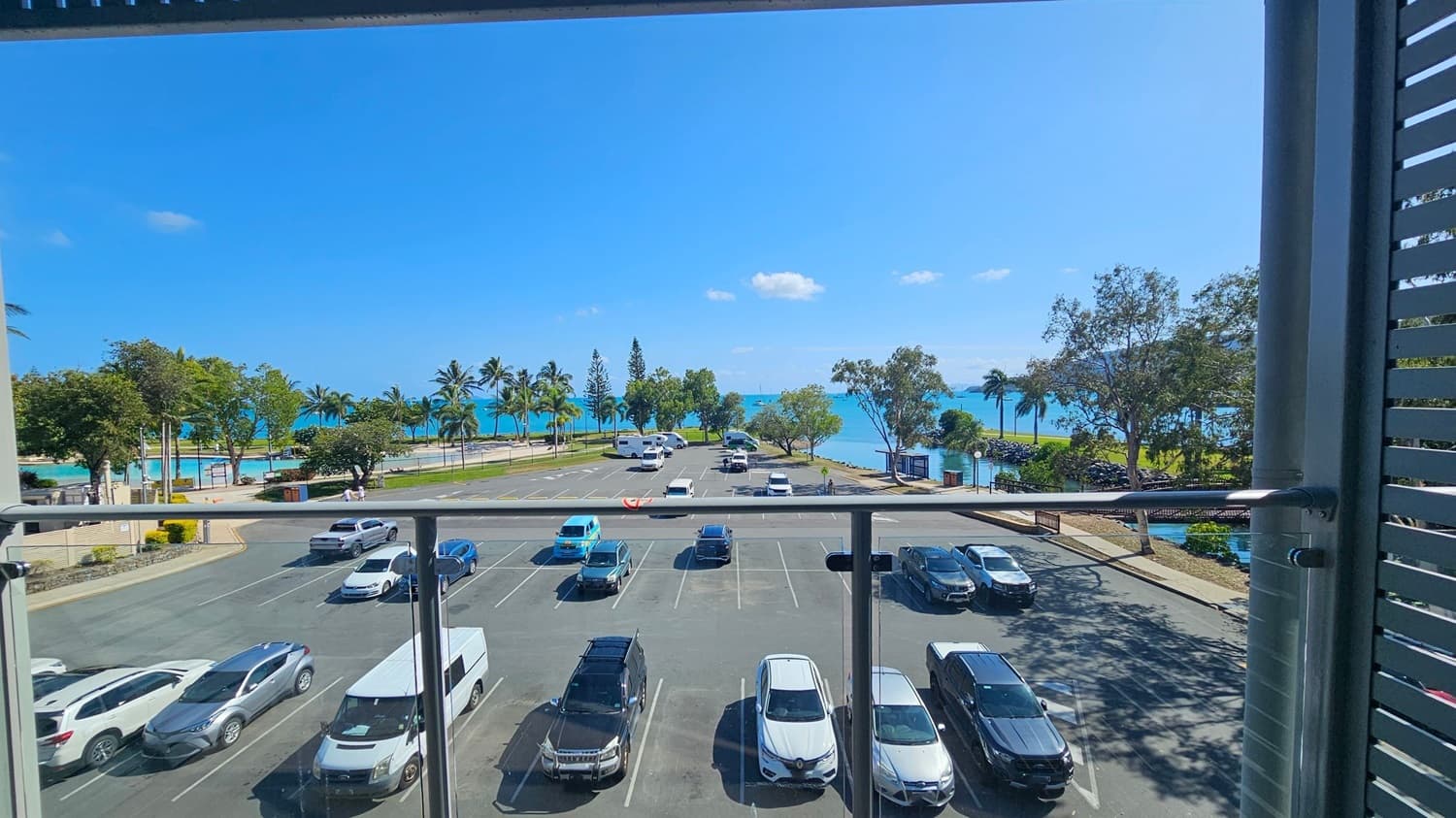 View from a balcony overlooking a car park with several vehicles, including cars and a van. Beyond the car park, a road lined with palm trees leads to a beach and the sea. The sea is visible in the background, with a few small boats on the water. Trees and greenery are scattered along the shoreline. The balcony has a glass railing, and the scene is framed by vertical slats on the right.