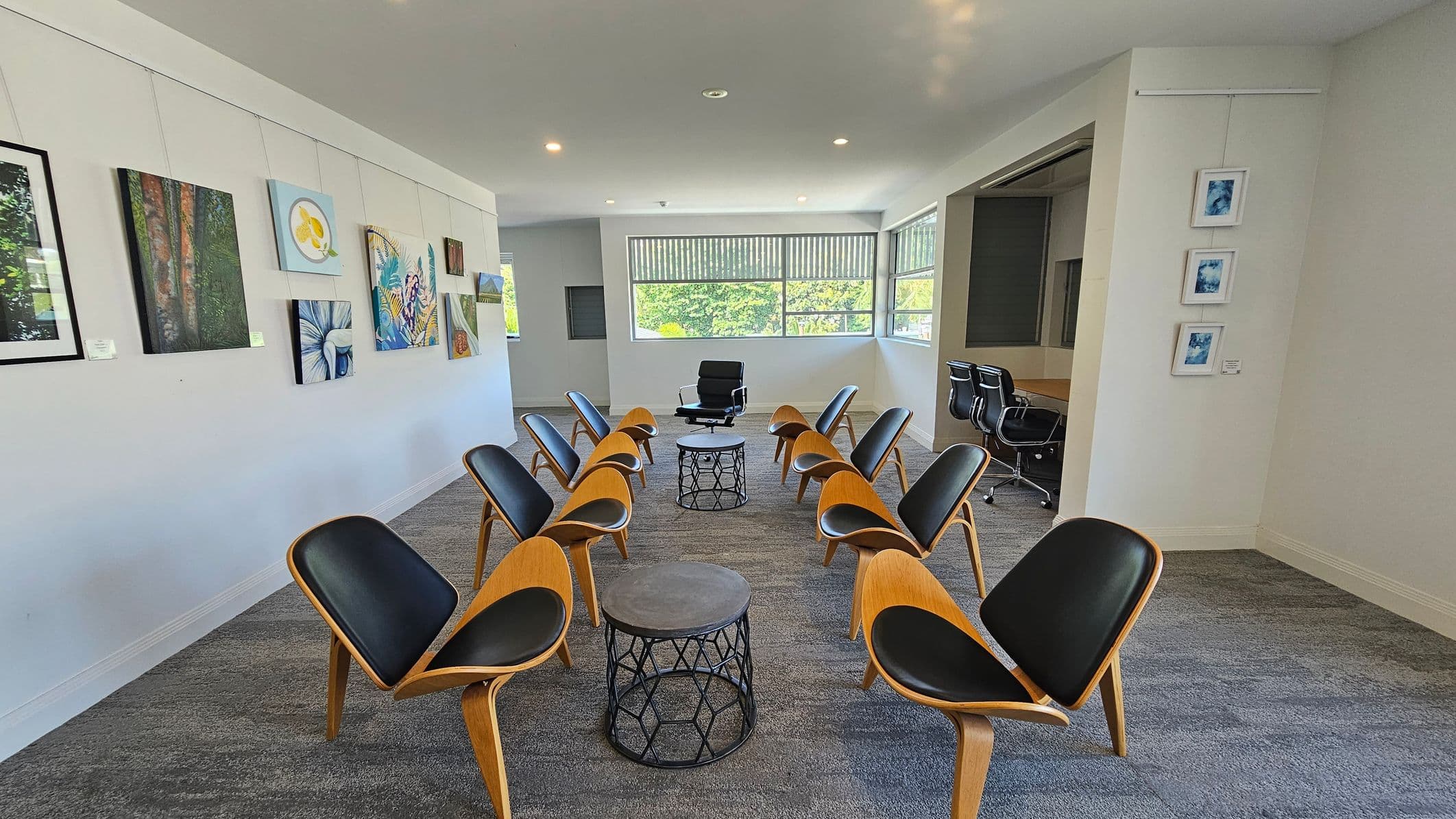 A room with a semi-circle arrangement of wooden chairs with black seats facing a single black chair and two round tables in the centre. The left wall displays various paintings, while the right wall has three small framed artworks. Large windows at the back let in natural light, and a conference table with office chairs is visible in an adjacent room. The floor is carpeted in a grey pattern, and the ceiling is plain with recessed lighting.