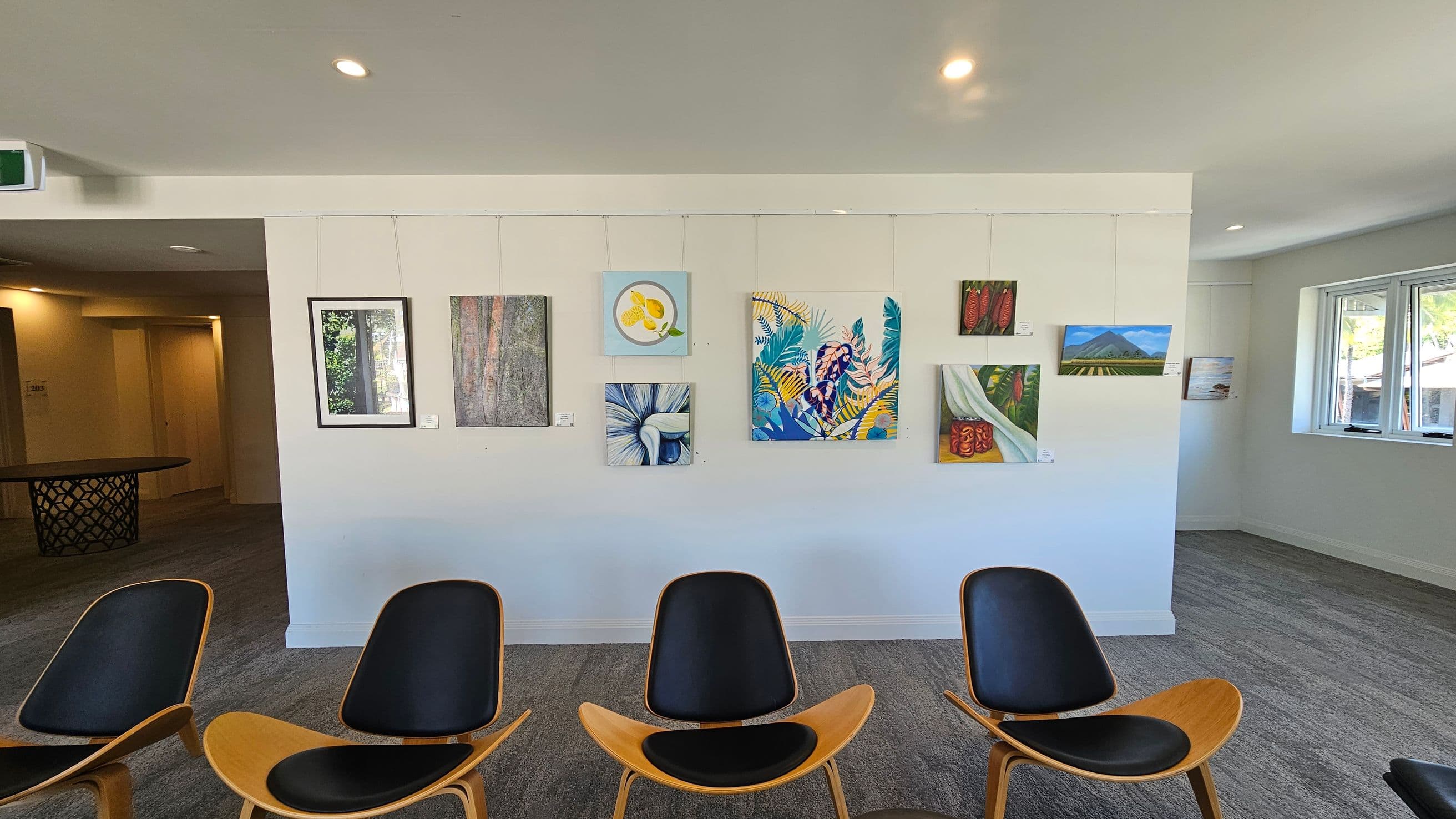A gallery wall with various paintings, including abstract and nature-themed artworks, is displayed. Below the paintings, four modern chairs with wooden frames and black seats are arranged in a row. To the right, there are windows allowing natural light into the room. The floor is carpeted in grey, and the ceiling is white with recessed lighting. On the left, a hallway leads to another area with a round table featuring a lattice design.