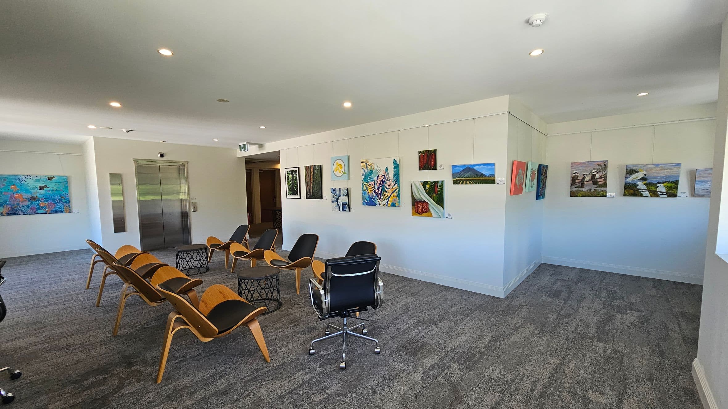 A modern gallery space with grey carpeted flooring and a white ceiling with recessed lighting. On the left, there is a wall with a colourful painting and a metal lift door. In the centre, several wooden chairs with black cushions are arranged around two small, round tables. On the right, a white wall displays various paintings, including landscapes and abstract art. A black office chair is positioned near the centre of the room.