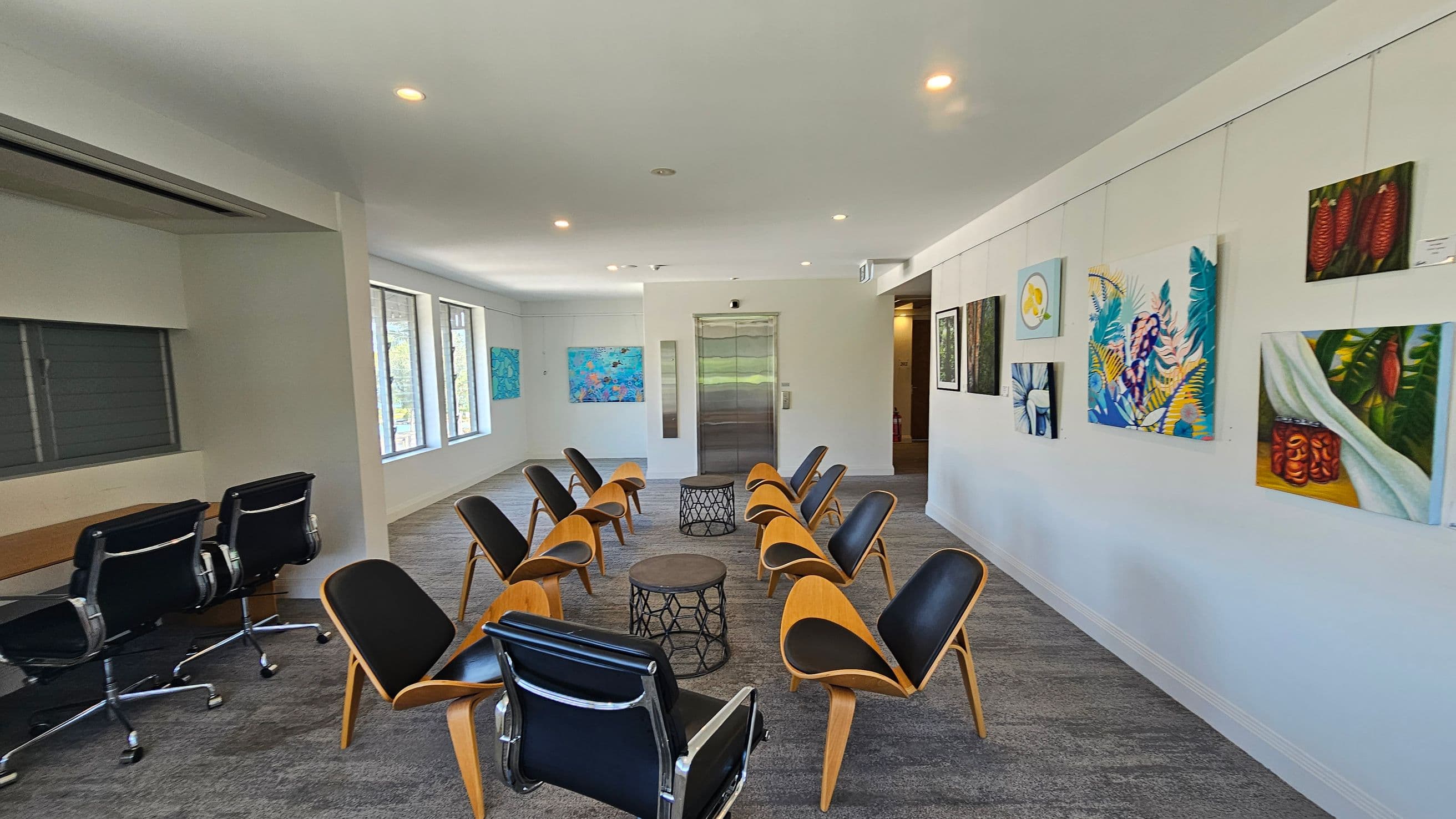 A modern room with grey carpeted flooring and a white ceiling with recessed lighting. In the centre, there are two circular tables surrounded by wooden chairs with black cushions. On the left, a row of black office chairs is positioned against a wall with windows. The right wall is adorned with colourful paintings. At the far end, an elevator is visible, flanked by more artwork.
