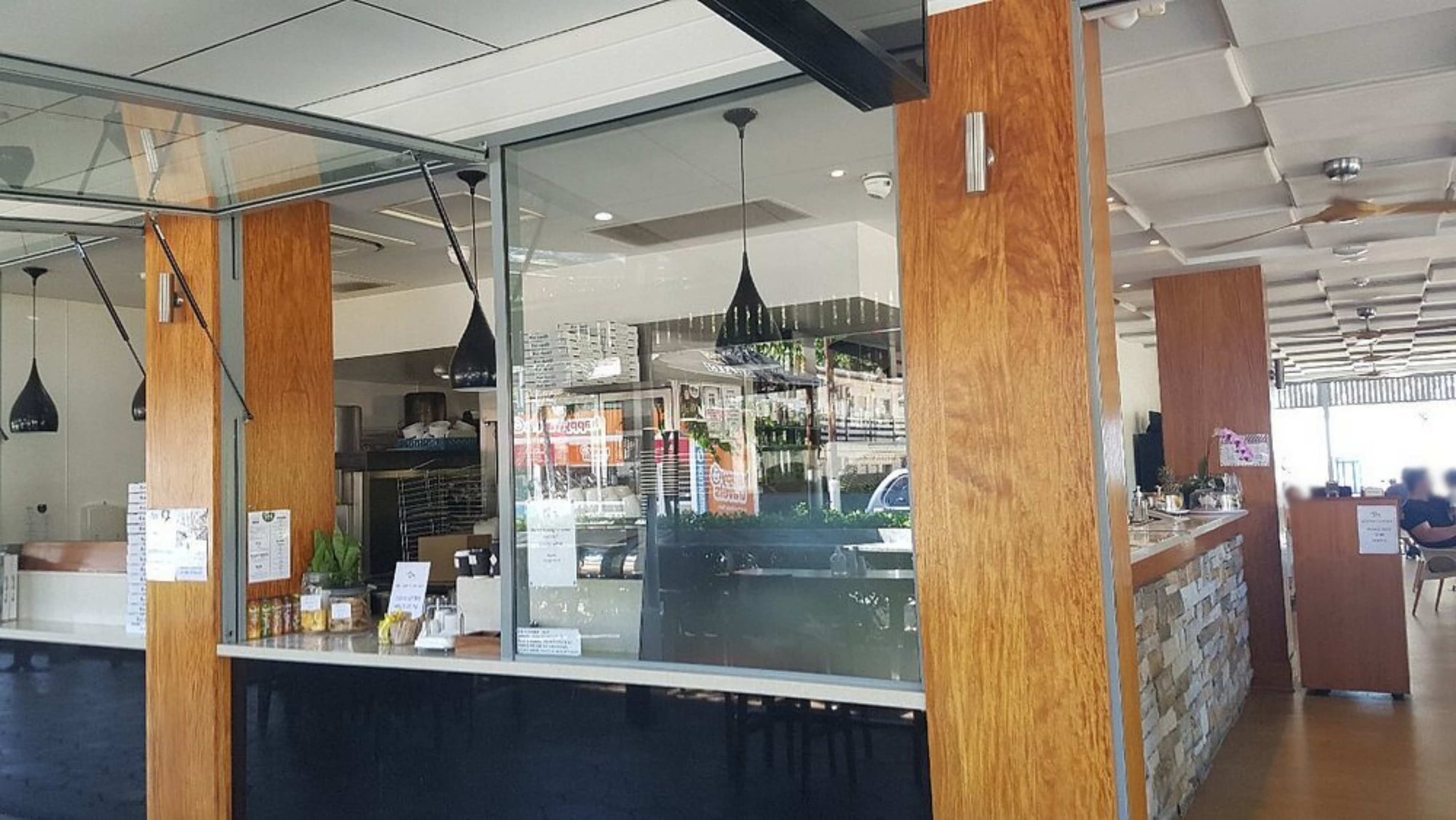 Wooden pillars are positioned on the left and right, framing glass windows. On the left, a counter displays small items, with black pendant lights hanging above. In the middle, glass panels reveal the interior, showing shelves and equipment. On the right, a stone-fronted counter runs along the wall, with tables and chairs in a dining area. The floor is light wood, and the ceiling has a grid pattern with recessed lights and ceiling fans.
