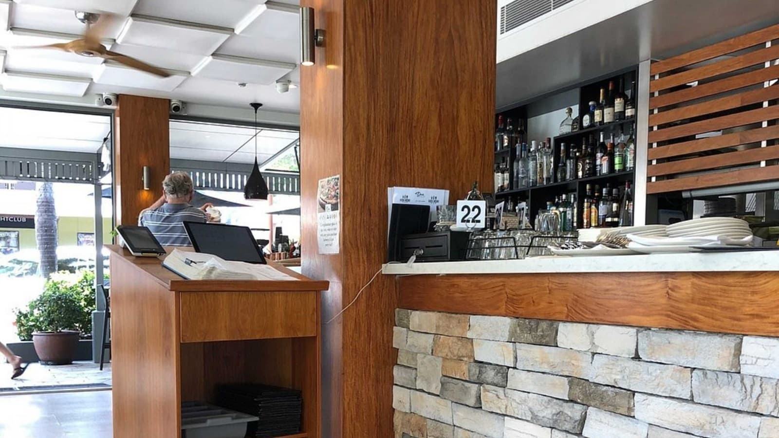 On the left, a wooden host counter with a register, papers, and a storage shelf below. A man in a striped shirt stands near glass doors, with outdoor seating visible beyond. In the middle, a wooden column extends from floor to ceiling. On the right, a stone-fronted counter topped with white marble holds plates, cutlery, and a numbered sign. Behind it, shelves with rows of bottles and glasses are framed by wooden slats. The floor is tiled, and the white ceiling has recessed panels.