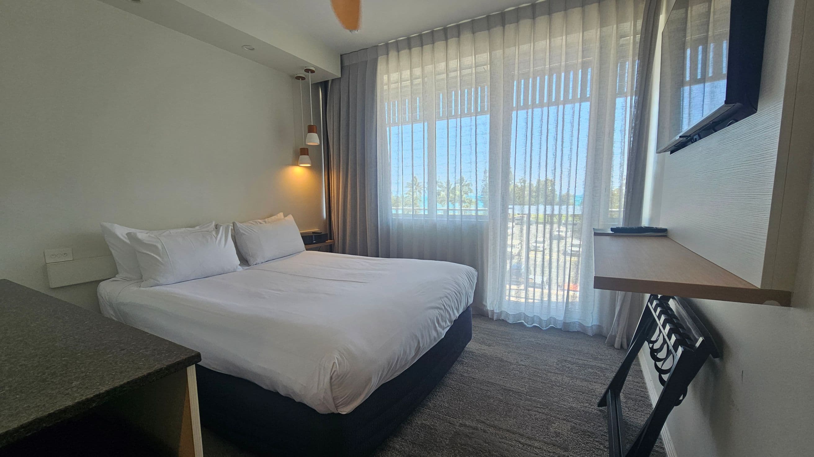 A hotel room with a large bed on the left, covered in white linens and pillows. Above the bed, a wall lamp provides light. On the right, a flat-screen TV is mounted on the wall above a small shelf. Below the shelf is a luggage rack. The room has a large window with sheer curtains, allowing natural light to filter in. The floor is carpeted in a neutral tone.