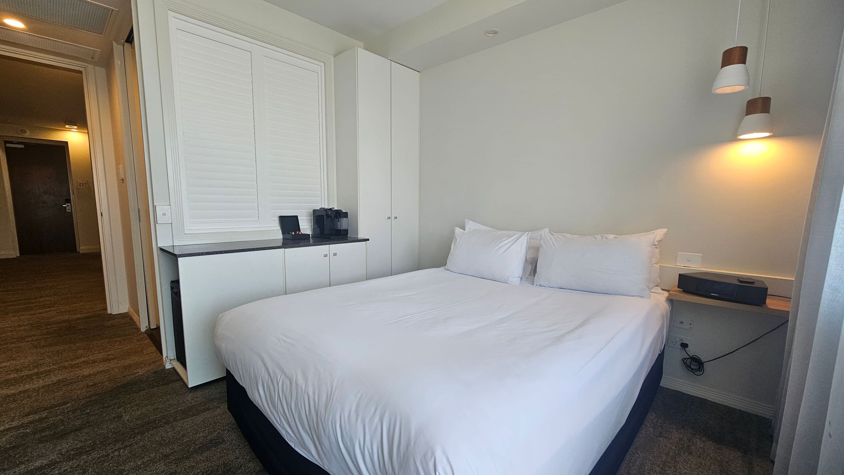 A hotel room with a neatly made double bed on the right, featuring white linens and two pillows. Above the bed, two pendant lights hang from the ceiling. On the left, there is a built-in cabinet with a countertop, holding a coffee machine and a tray. The floor is carpeted in a mix of green and brown tones. An open door leads to a hallway with a visible room number, 1217, on a dark door. The walls are light-coloured, and the ceiling is plain with recessed lighting.