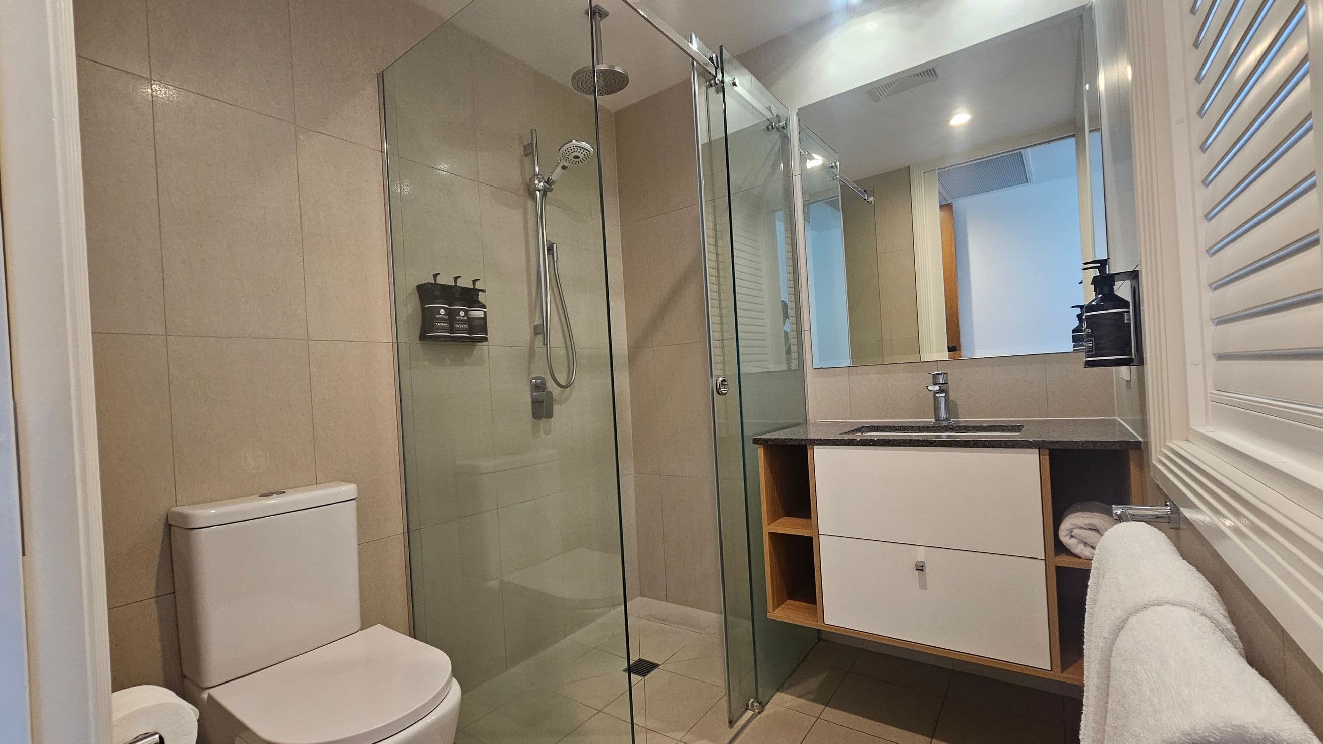 Modern bathroom with beige tiled walls and floor. On the left, a white toilet is positioned next to a glass-enclosed shower with a rainfall showerhead and mounted toiletries. On the right, a vanity with a black countertop holds a sink, with a large mirror above. Below the sink, there are open shelves and a towel rack with a white towel. The ceiling has recessed lighting, and a louvred panel is visible on the right wall.