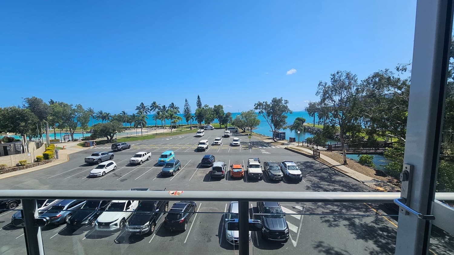 A view from a balcony overlooking a car park with several vehicles parked in rows. Beyond the car park, there are palm trees and a grassy area leading to a turquoise sea. On the right, a small bridge crosses over a narrow waterway. The scene is framed by a glass railing in the foreground.