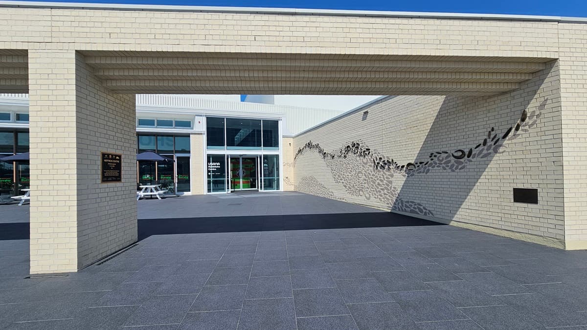 The building has a generous front with a brick wall facade featuring intricate design. A plaque displaying facility information is mounted on the left wall. Behind it is a two-story modern structure with glass walls and corrugated roofing. Tables and chairs are arranged under large umbrella shades.