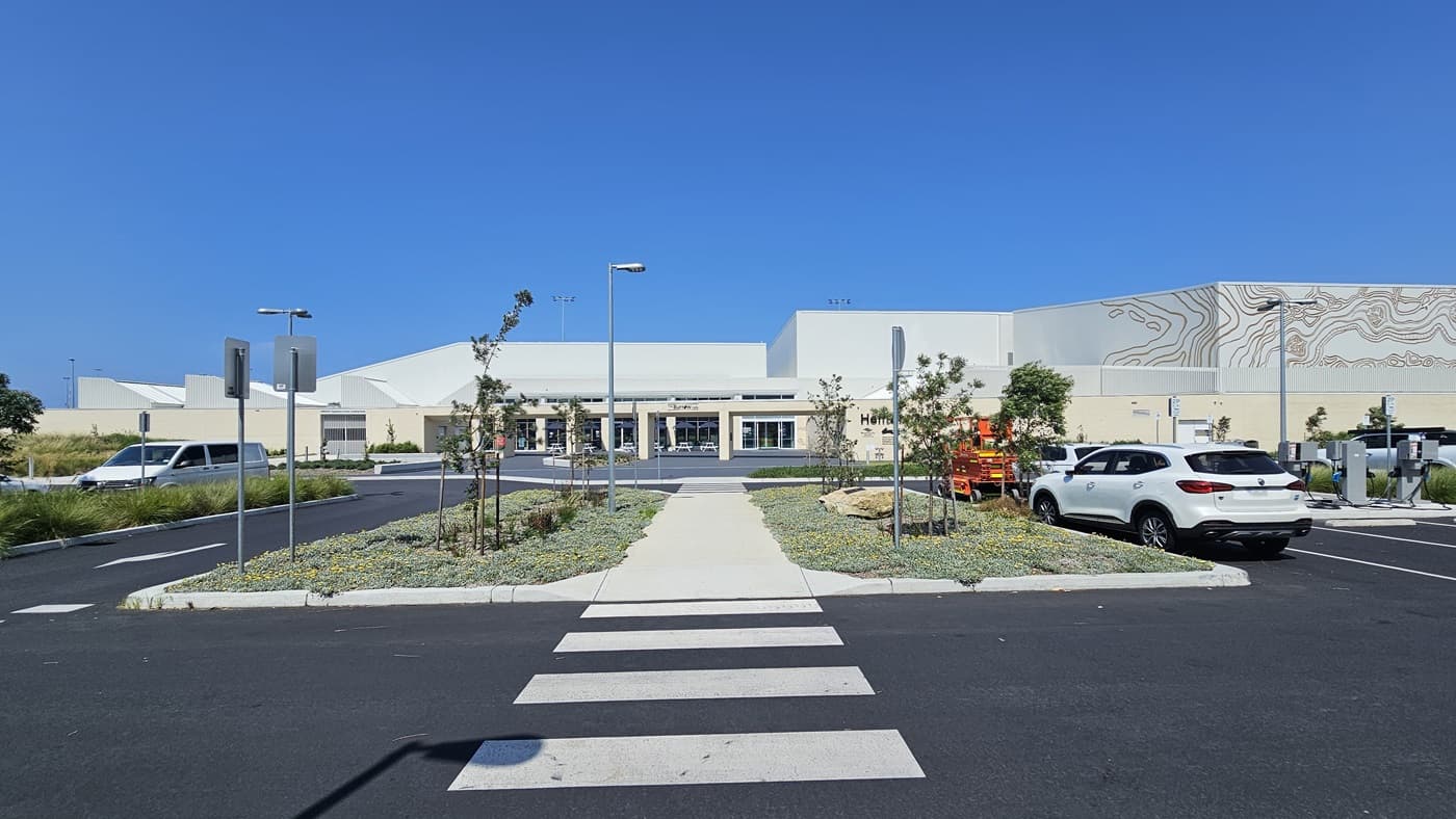 The building displays a generous front with a brick wall facade featuring intricate design and the facility name. Next to it is the car park, occupied by a few cars. In the middle, there's a landscape of plants and small trees. A pedestrian crossing and concrete pathway run between the two landscapes, extending towards the building. Street signs and street lights are visible.
