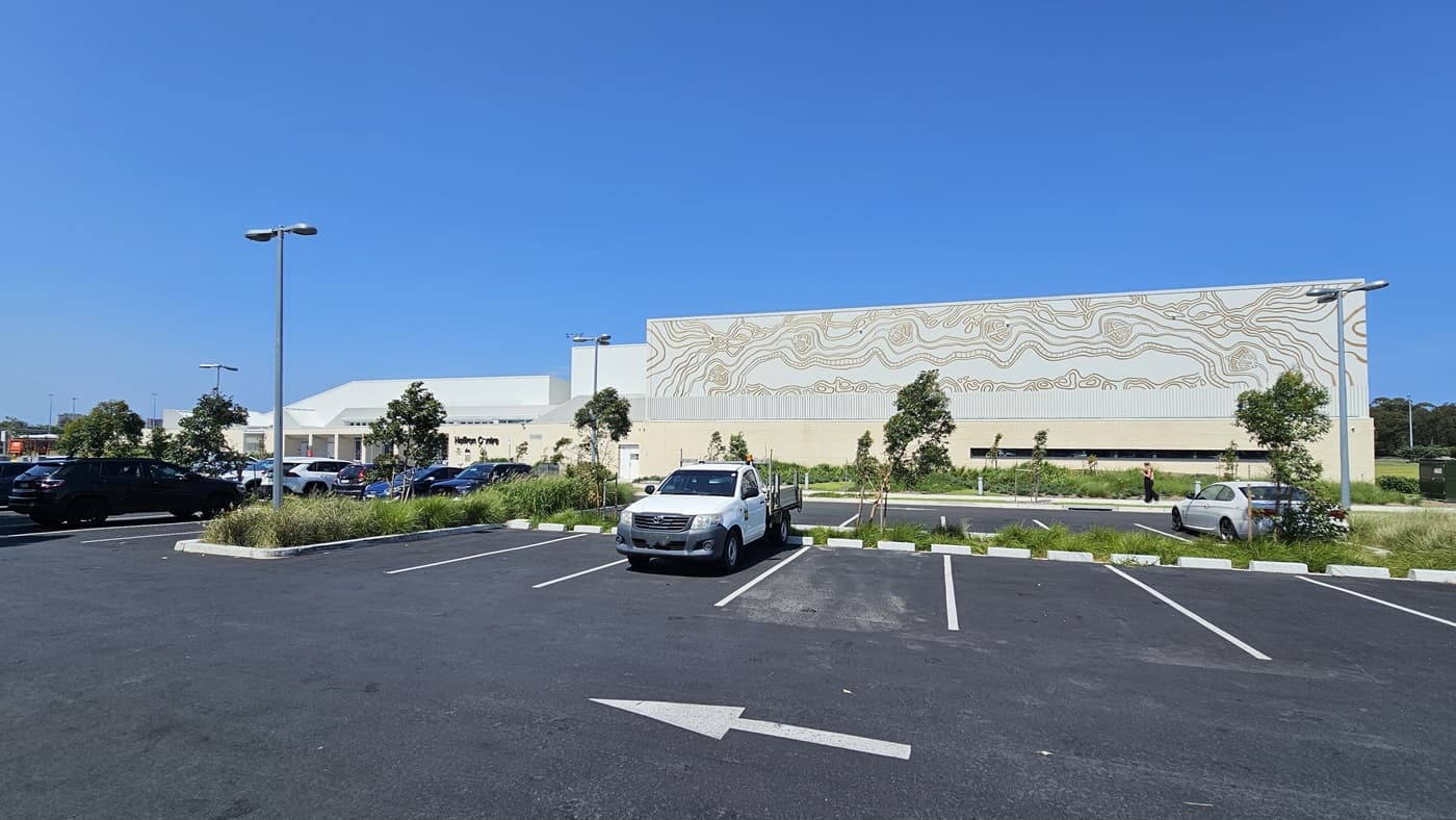 Next to the facility is the car park, featuring line markings and occupied by a few cars. On the left, there's a concrete plant box with trees and grass. Adjacent to it is the building, presenting a brick wall facade with intricate design and the facility name. Street lights are visible.