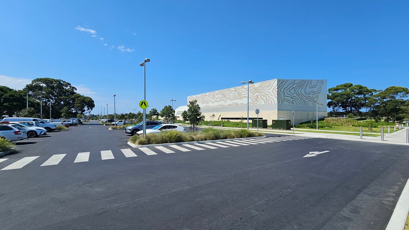 A pedestrian lane runs parallel to concrete plant box landscapes. Street signs and street lights are visible. To the left, the car park is occupied by cars of various shades and models. To the right, the building displays a brick wall facade with intricate designs. Concrete bollards and trees are visible on the right.