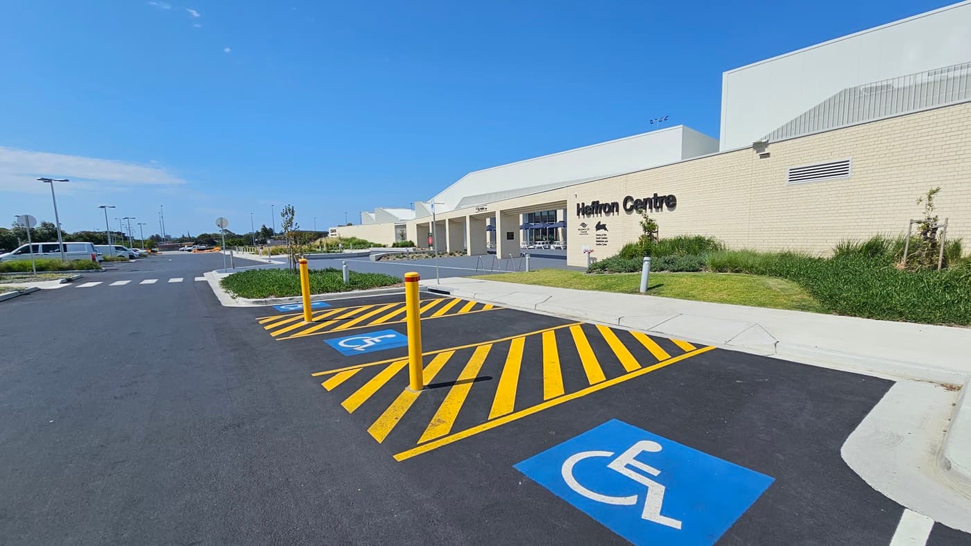 The building features a spacious front with a brick wall facade, showcasing intricate design and the facility name. To the right, there are accessible car parking spaces marked by blue wheelchair signs, yellow bollards, and yellow floor markings. In the middle, a pedestrian lane and concrete pathway run between two landscapes, extending towards the building. Street signs and street lights are visible.