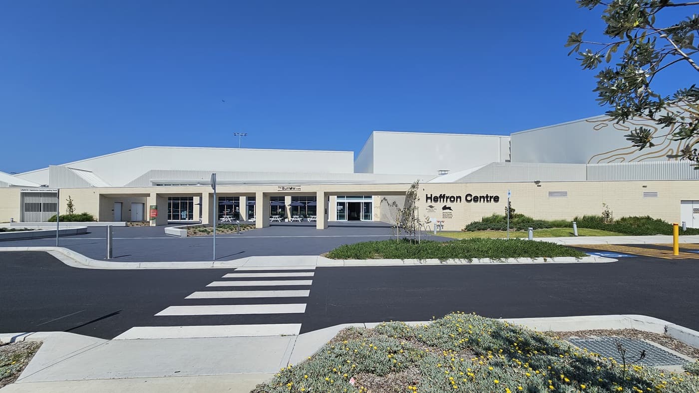 The building features a spacious front with a brick wall facade, displaying the facility's name and its partners. On the right, there's a landscape with grass and ornamental plants. Next to the wall is the accessible parking area, equipped with signs and concrete bollards. Behind it stands a two-story modern structure with glass walls and corrugated roofing. Tables and chairs are arranged under large umbrella shades. A pedestrian crossing extends from the car park.