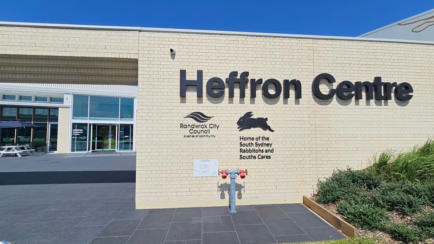 The building features a spacious front with a brick wall facade, showcasing the facility's name and its partners. On the right, there is a landscape of grass, ornamental plants, and a fire hydrant. Behind it is a two-story modern structure with glass walls and corrugated roofing. Tables and chairs are arranged under large umbrella shades. The floor is tiled in grey.