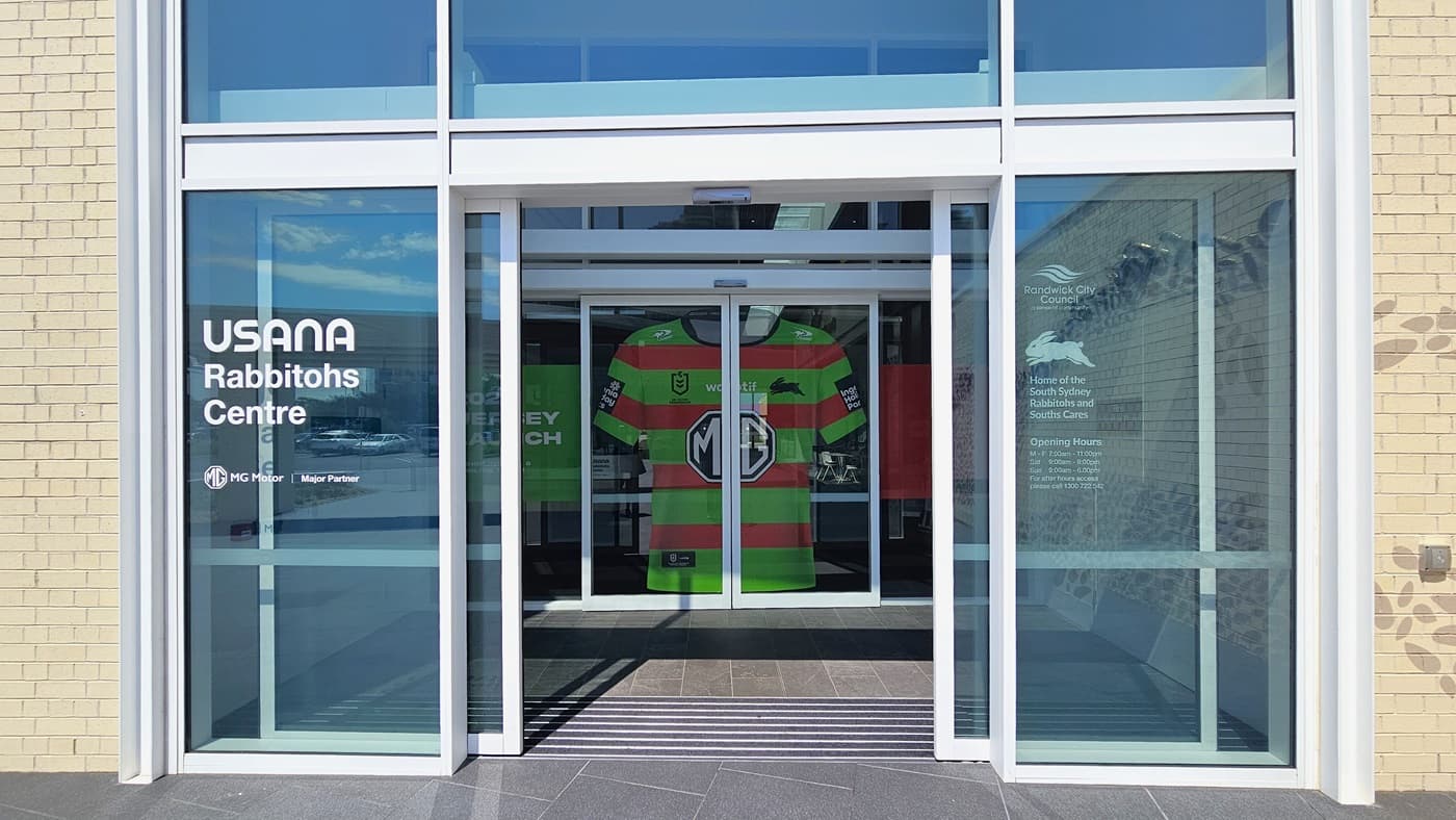 Access to the Heffron Centre is through a set of double automatic glass doors equipped with non-slip mats. Stickers displaying the facility's partners are on the left, and the store hours are on the right. The second glass door features a large orange and green t-shirt sticker, spanning the entire door and splitting when opened. The outer wall features a cream brick design.