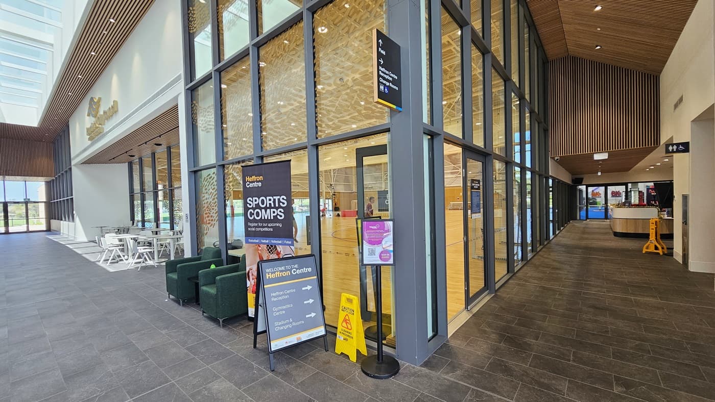 To the left, tables and chairs are arranged. The glass wall features a golden logo and small leaf-like designs in gold colors, with free-standing banners positioned on the wall. To the right is a corridor leading to the Heffron Centre reception area. The floors have matte grey tiles, and the ceiling features intricately designed wooden panels with LED lights. Further right, the reception desk and the gymnastics area are visible.