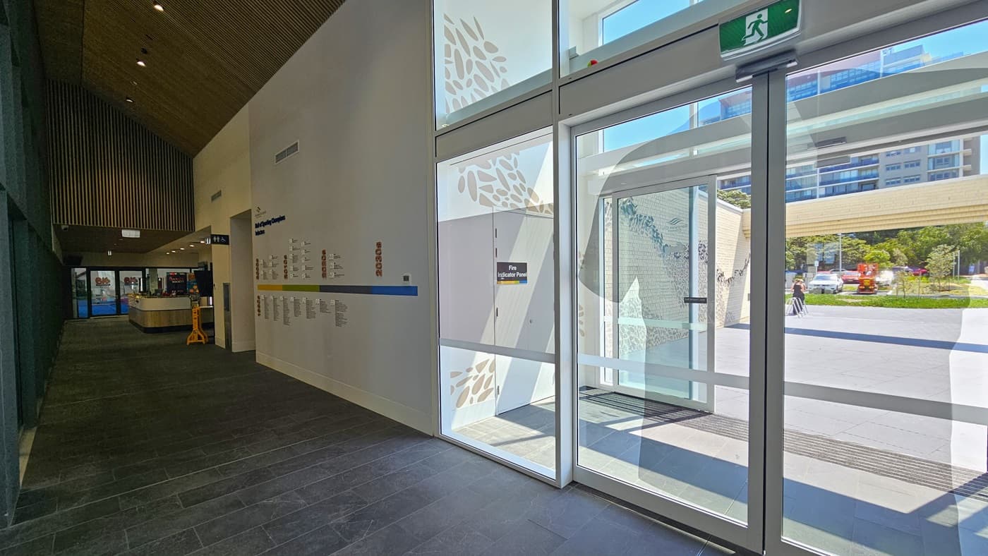 The corridor leads to the Heffron Centre reception desk. The right wall features a 'Hall of Sporting Champion Inductees.' Straight ahead is the reception desk and the gymnastic area. To the right is the entrance to the building featuring double automatic glass doors. The wall outside features intricate designs. Outside, the car park, tall commercial buildings, and trees are visible.