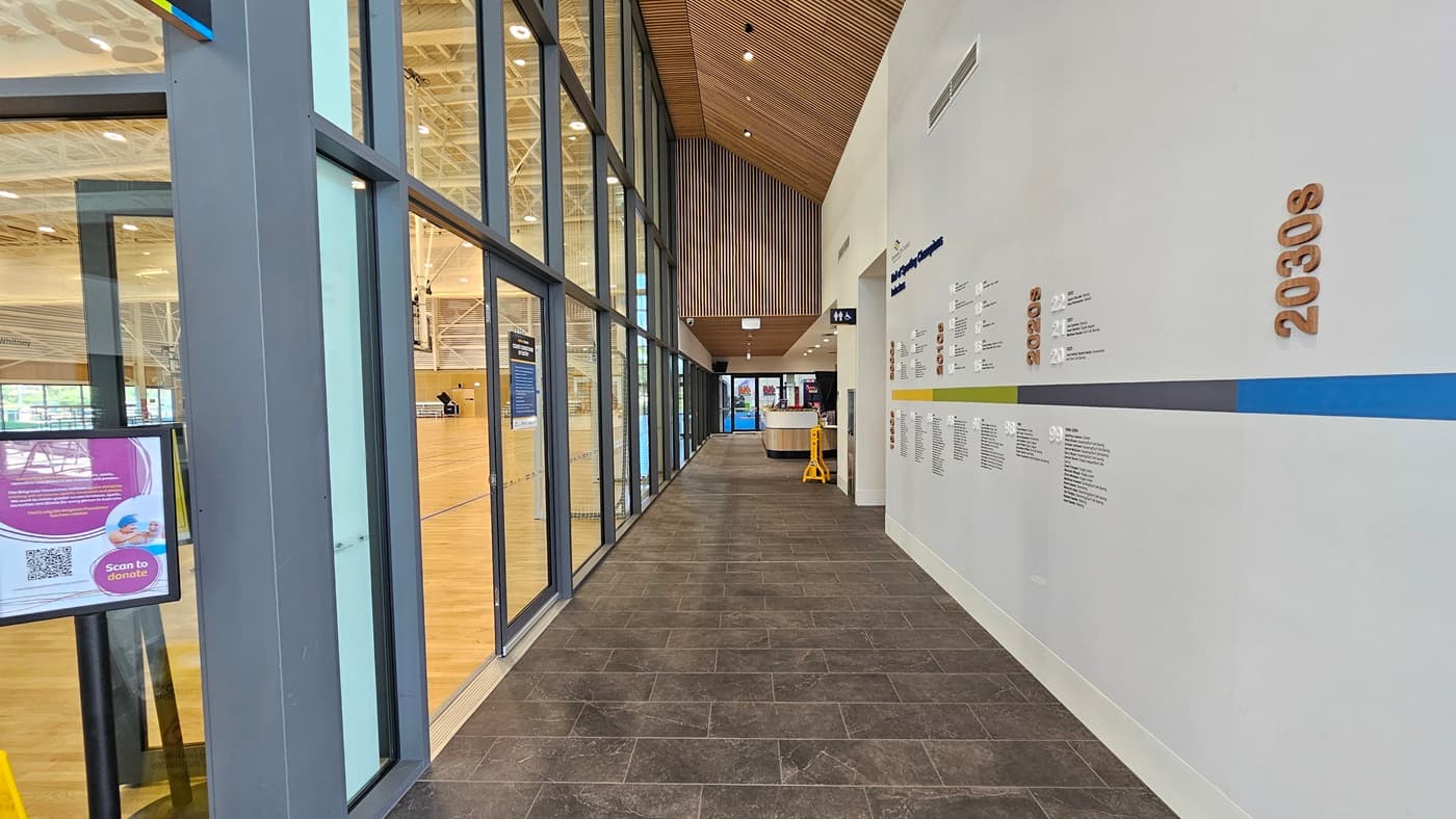 The corridor directs to the Heffron Centre reception desk. The right wall features a 'Hall of Sporting Champion Inductees.' Straight ahead is the reception desk and the gymnastics area. To the left is the sports stadium with timber floors and an industrial ceiling. Posters are affixed to the glass wall. The corridor has grey tiles, and the ceiling features an intricate wooden design.