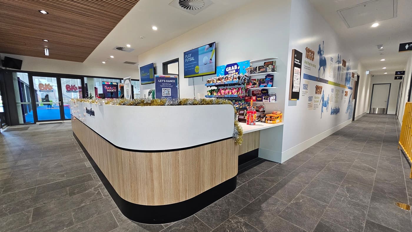 The reception desk displays a white and timber design front with food items, banners, posters, and garlands. Two flat-screen TVs are mounted on the wall. The right corridor has a white wall painted with historical facts, and accessible bathroom signage is mounted on the walls to the right. To the left is the gymnastics area. The corridor has grey tiles, and the ceiling features an intricate wooden design.