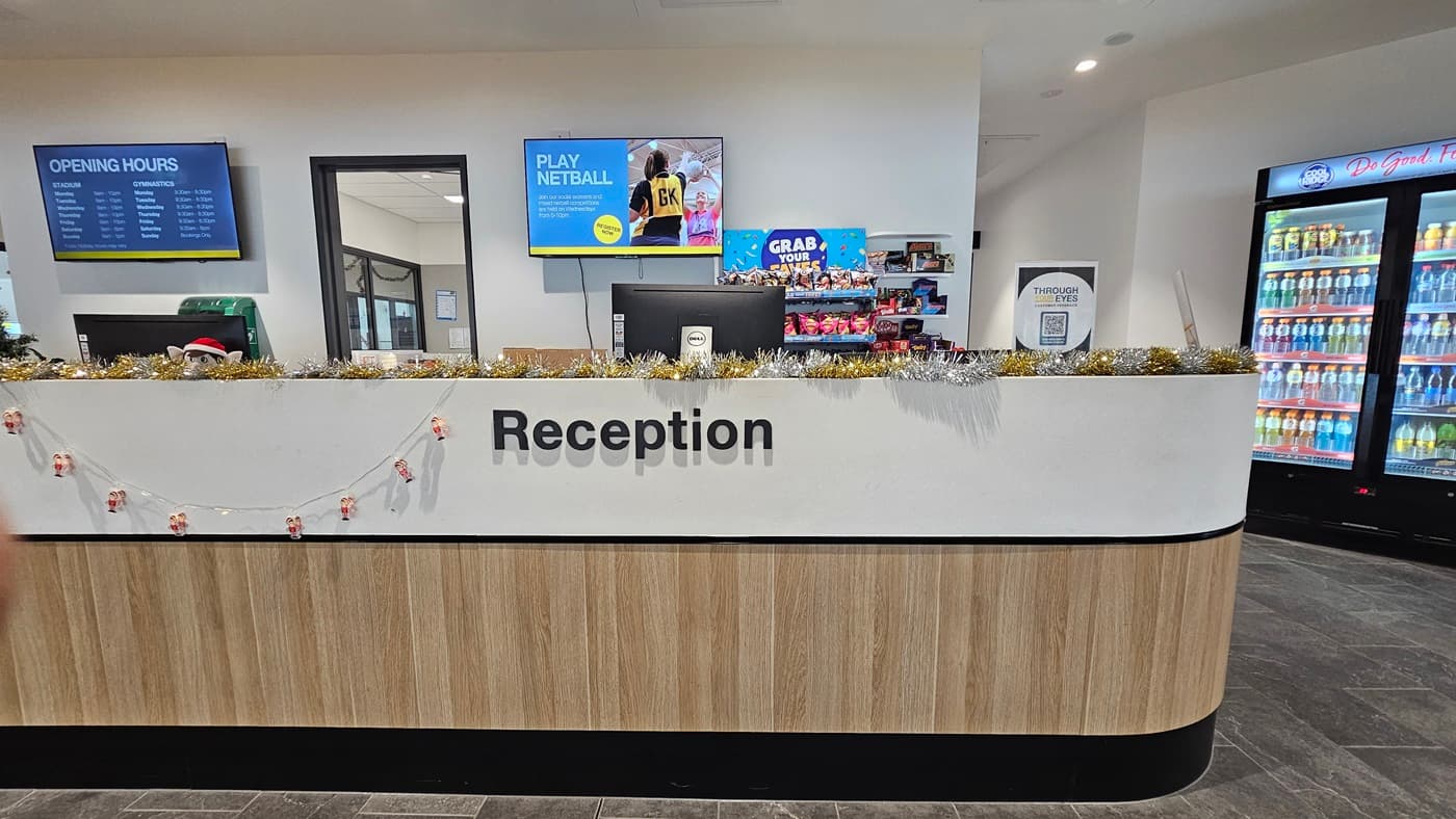 The reception desk has a white and timber design front, displaying food items, banners, posters, and garlands. Two computers are placed on top of the desk. In the middle, a door leads to the staff office. Two flat-screen TVs are mounted on the wall. To the right, a vending machine offers cold drinks. The floor has grey tiles, and the ceiling is all white with LED lights.