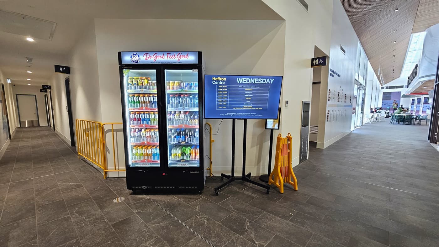 In the middle is a vending machine offering cold drinks. Beside it is a flat-screen TV, and collapsible barricades are also visible. Adjacent are the accessible toilets and a drinking fountain. The corridor to the right leads to a seating area where tables and chairs are arranged. The corridor to the left leads to male change rooms. The floor has grey tiles, and the ceiling is all white with LED lights.