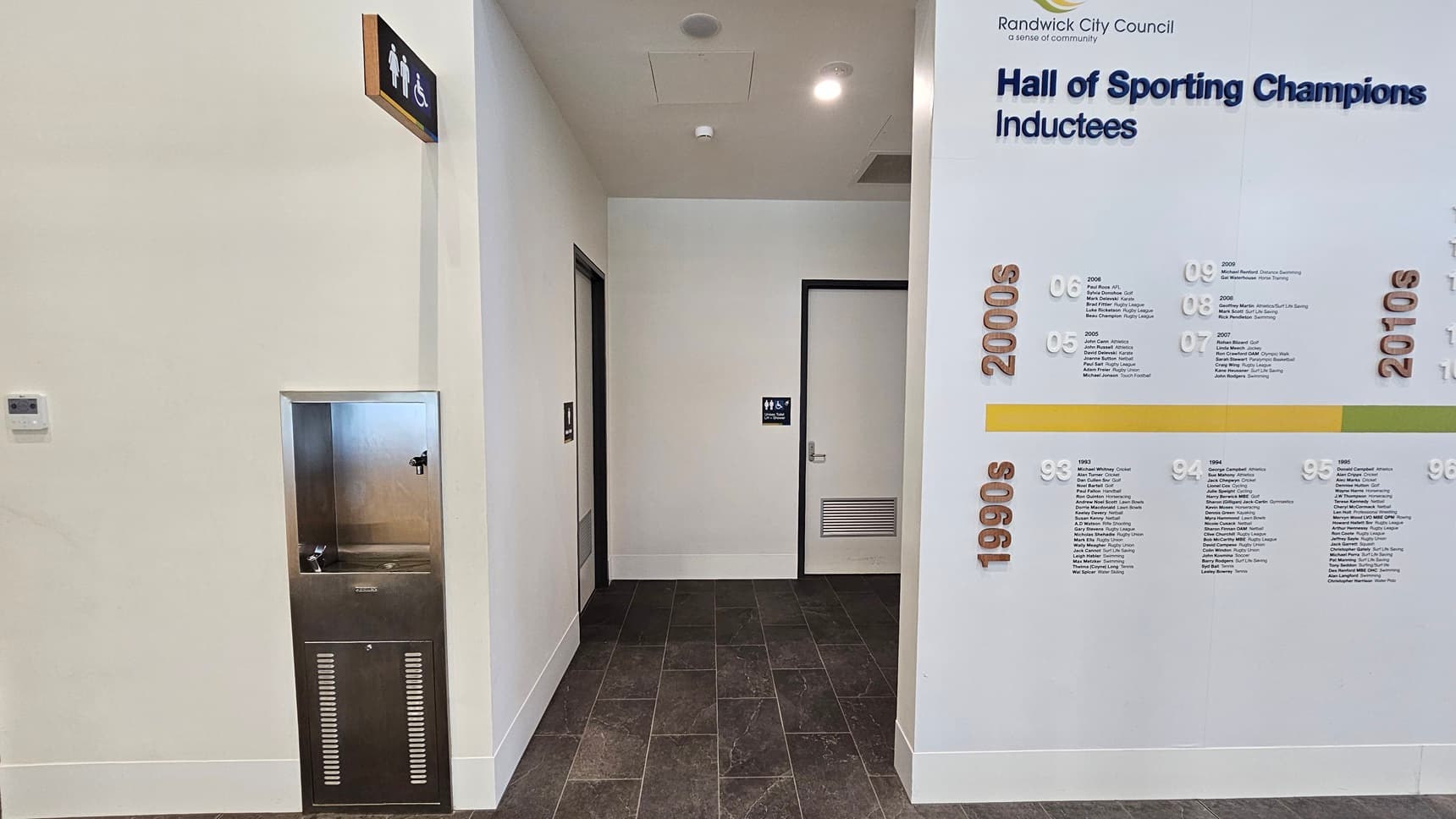 To the left is a drinking fountain and water refilling station. A corridor leads to accessible bathrooms, each door marked with signage indicating gender access. The right wall features a "Hall of Sporting Champion Inductees." The floor has grey tiles, and the ceiling is all white with LED lights.