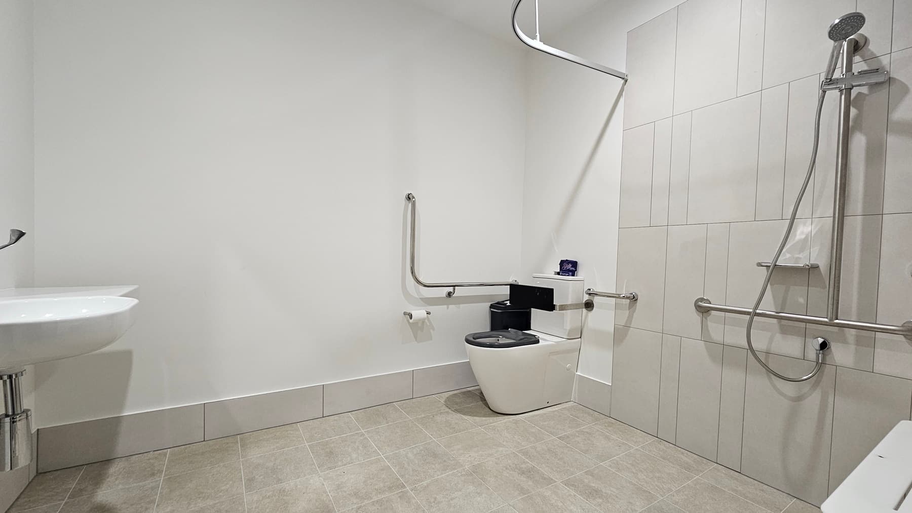 Inside the accessible bathroom, to the right corner is a toilet bowl equipped with hand grabs, accompanied by a rubbish bin. Adjacent is a shower with hand grabs and a placeholder for a shower curtain. A foldable bench chair is positioned to its right. A washbasin and a tissue dispenser are mounted on the left wall. The floor features grey tiles, and the wall is all white.