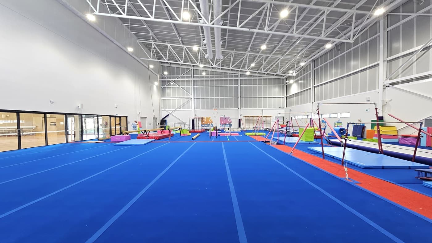 The floor is made of bright blue bouncy foam. The middle section is a spacious blank space. Further in, various gymnastics equipment such as balance beams, single bar trainers, horizontal bars, vault springboards, mats, and pommel horses are available. The high ceiling features an industrial look with exposed white metal beams. The left section is a glass wall that offers a view of the Sports Stadium.