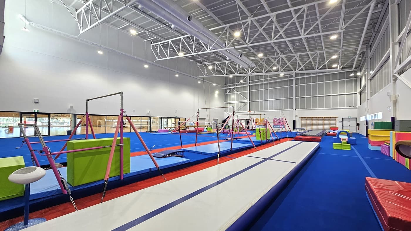 The floor is made of bright blue bouncy foam. On the left is a spacious blank space. To the right, various gymnastics equipment such as balance beams, single bar trainers, horizontal bars, vault springboards, mats, and pommel horses are available. The high ceiling features an industrial look with exposed white metal beams. The left section is a glass wall that offers a view of the Sports Stadium.