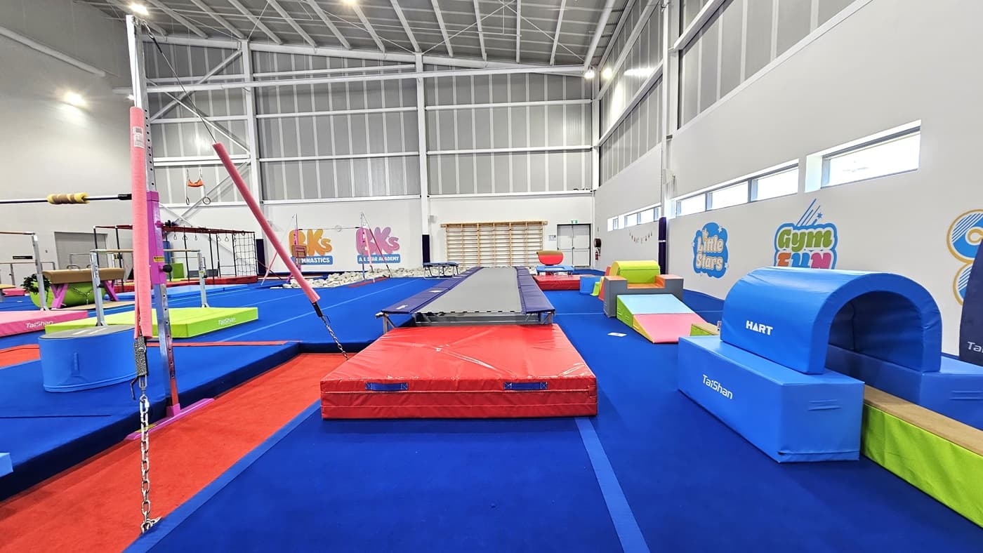 The floor is made of blue bouncy foam. The left and right walls feature white with paintings of the facility's name. Various gymnastics equipment such as balance beams, single bar trainers, horizontal bars, vault springboards, mats, and pommel horses are available. Brightly colored trampolines and tumbling blocks are also present. The high ceiling features an industrial look with exposed white metal beams.