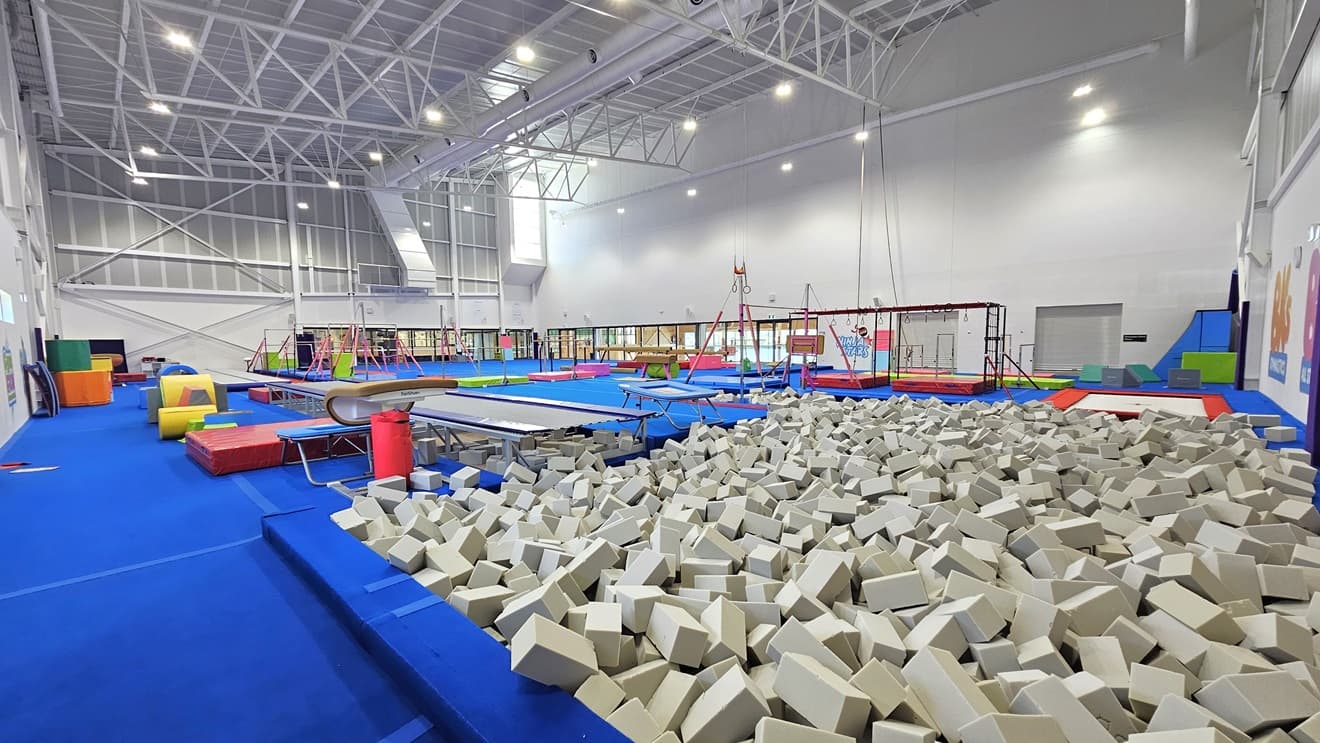 The floor is made of blue bouncy foam. The left and right walls feature white with paintings of the facility's name. Various gymnastics equipment such as balance beams, single bar trainers, horizontal bars, vault springboards, mats, and pommel horses are available. Brightly colored trampolines and tumbling blocks are also present. A foam pit with grey foams is positioned near the wall on the right. The high ceiling features an industrial look with exposed white metal beams.